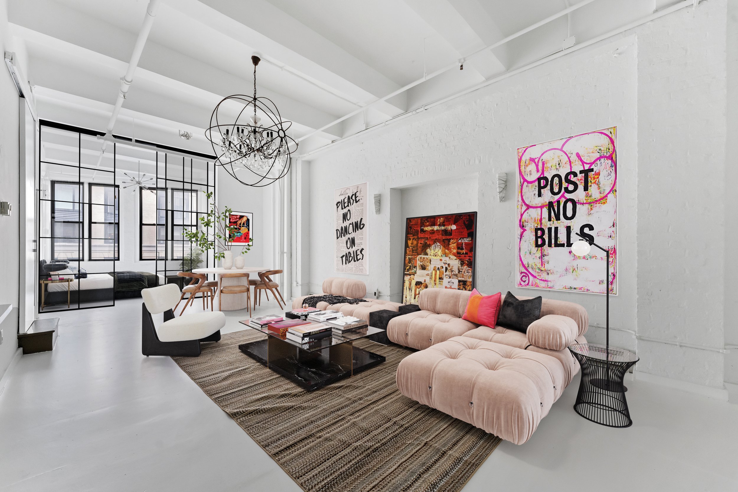 Modern, bright living room with a pink velvet sectional sofa, colorful throw pillows, and a black wire side table with a white lamp. Artwork on white brick walls, a black coffee table with books, and a striped rug. Dining area with a round wooden tab