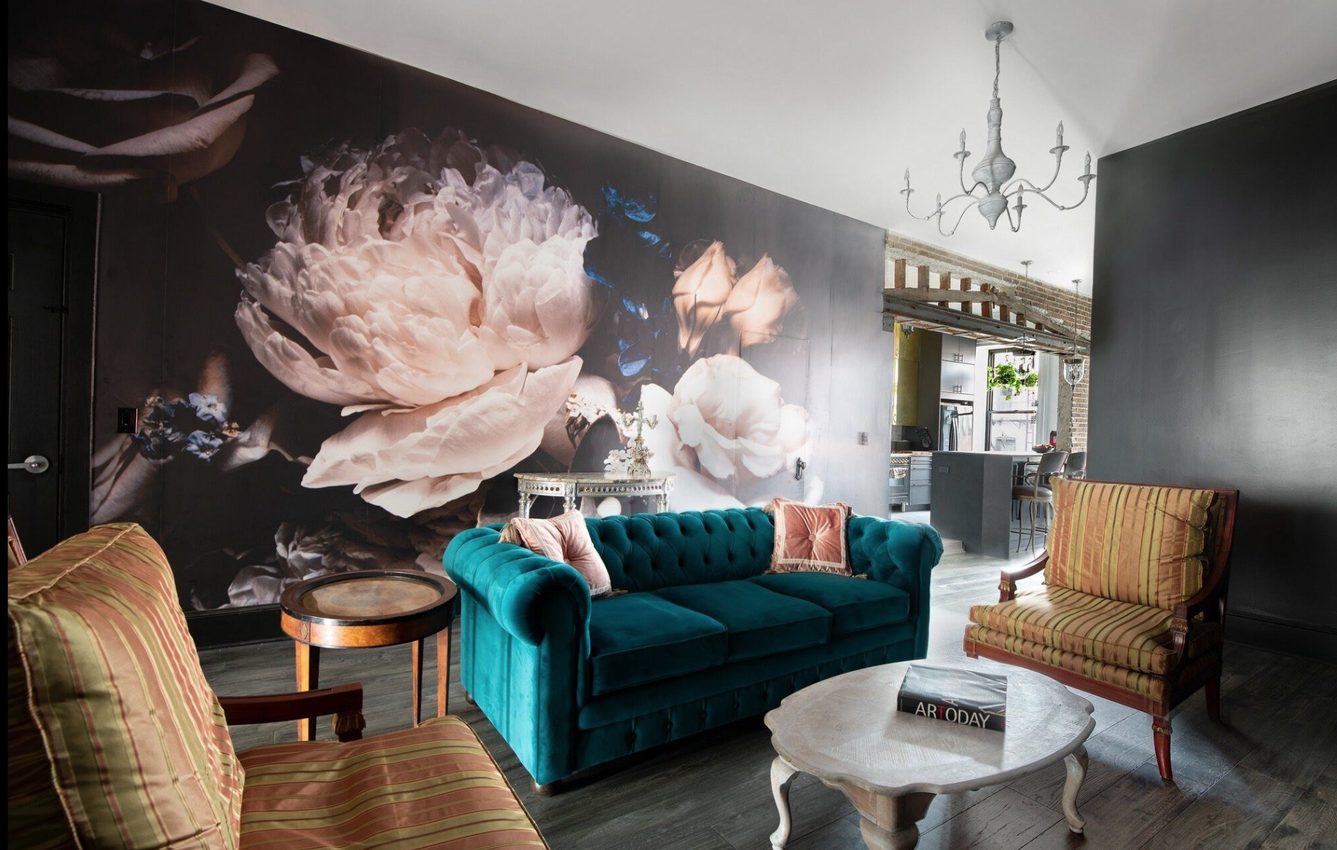 Living room with a dark floral feature wall, velvet teal sofa with cushions, a marble coffee table, and striped armchairs, with a chandelier and visible kitchen in the background.