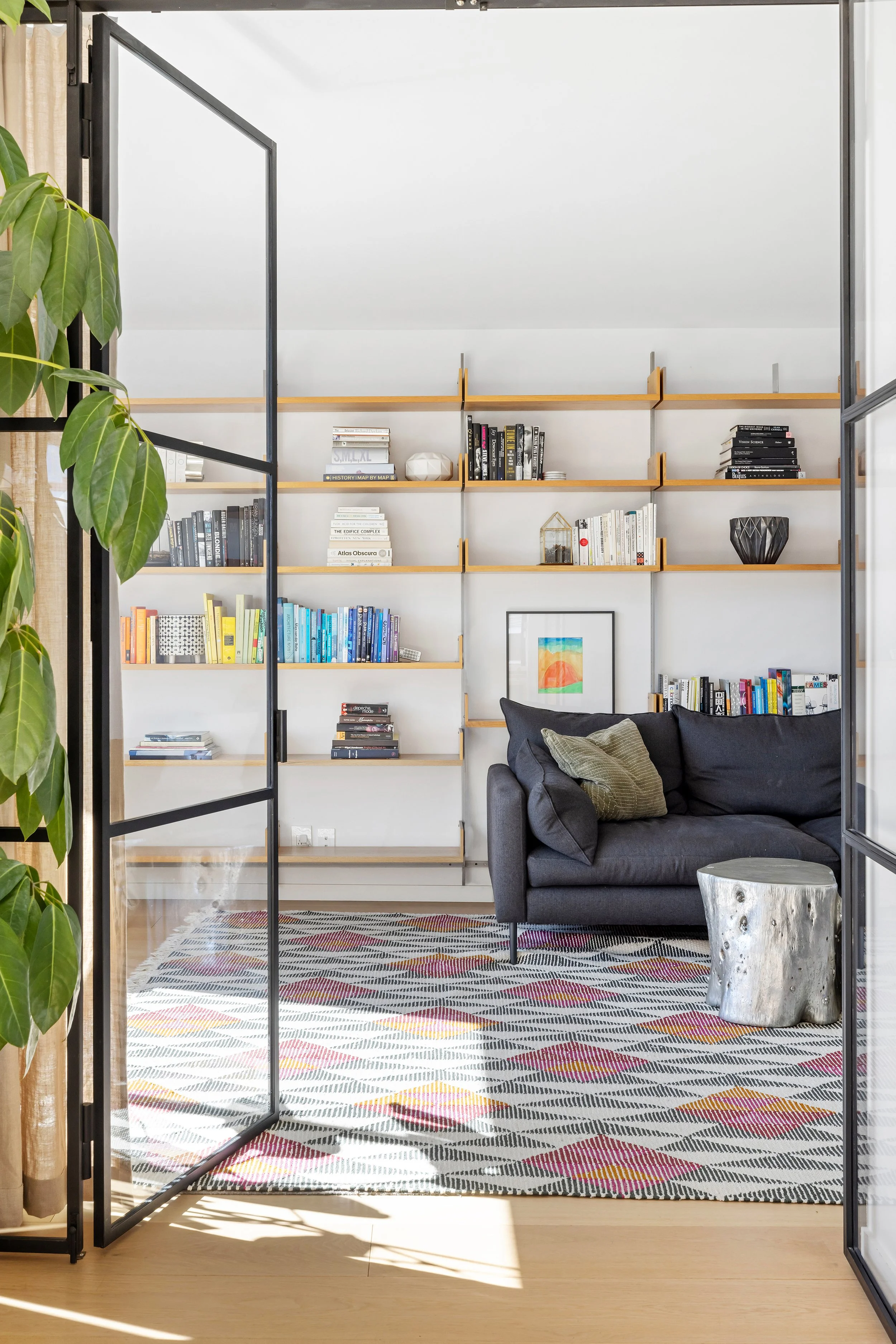 A cozy living room with a dark gray sofa, a colorful patterned rug, a white wall with wooden shelves filled with books and decorative items, and a tree with large green leaves on the left side. The room is viewed through a set of open glass and black