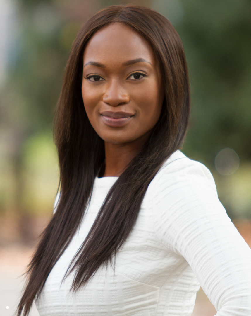 A confident African American woman with straight, shoulder-length hair, wearing a white top, standing outdoors with a blurred background of greenery.