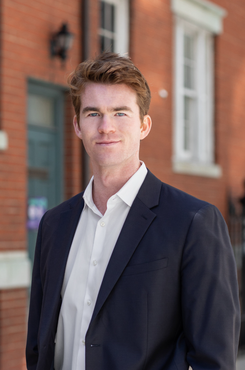 A young man with light skin, red hair, and blue eyes wearing a white shirt and a dark blue blazer standing outdoors in front of a brick building with windows.