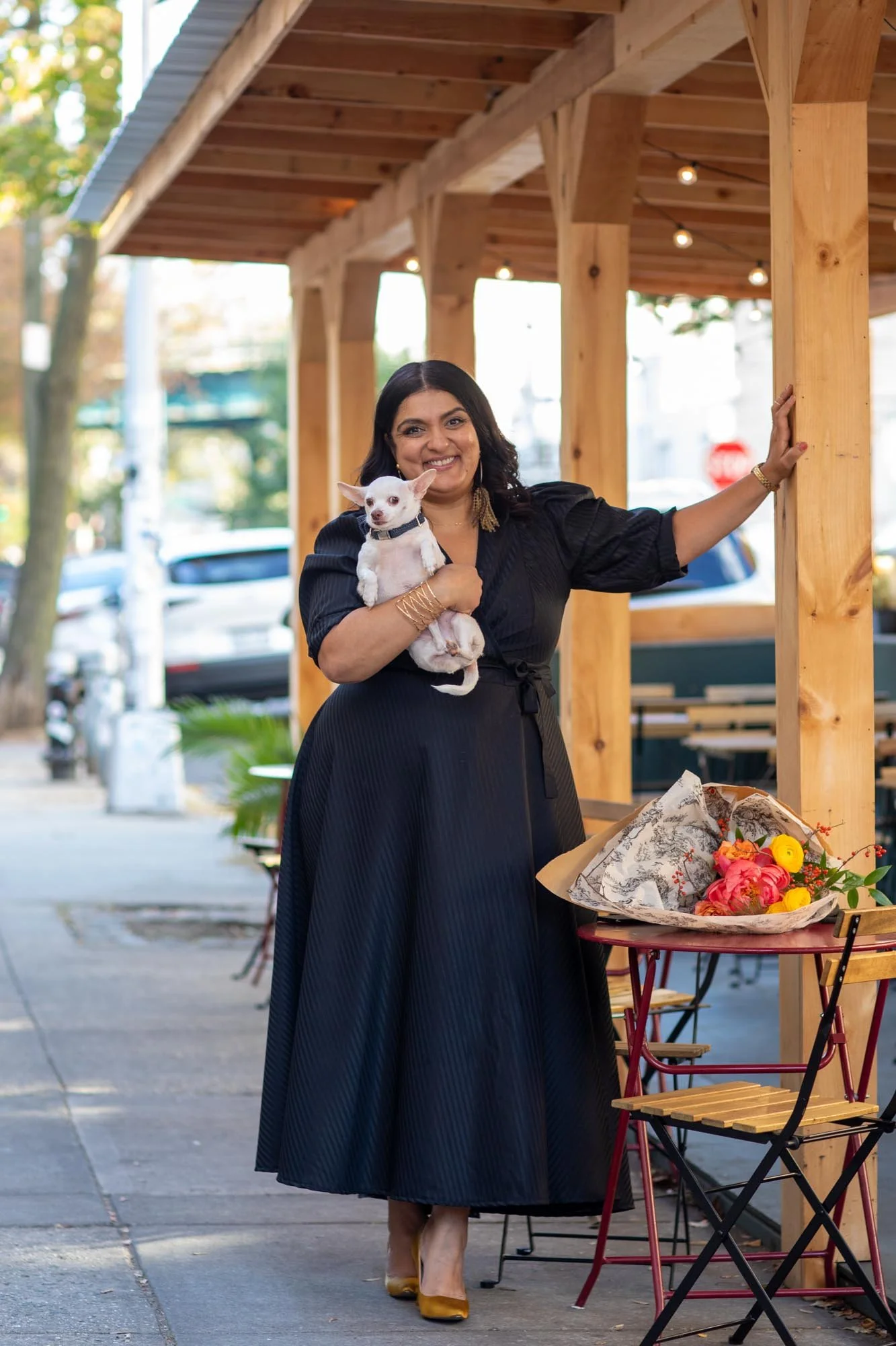 A woman wearing a black dress and yellow heels standing on a sidewalk outside a wooden structure, holding a small white dog, with a bouquet of flowers placed on a small table nearby.