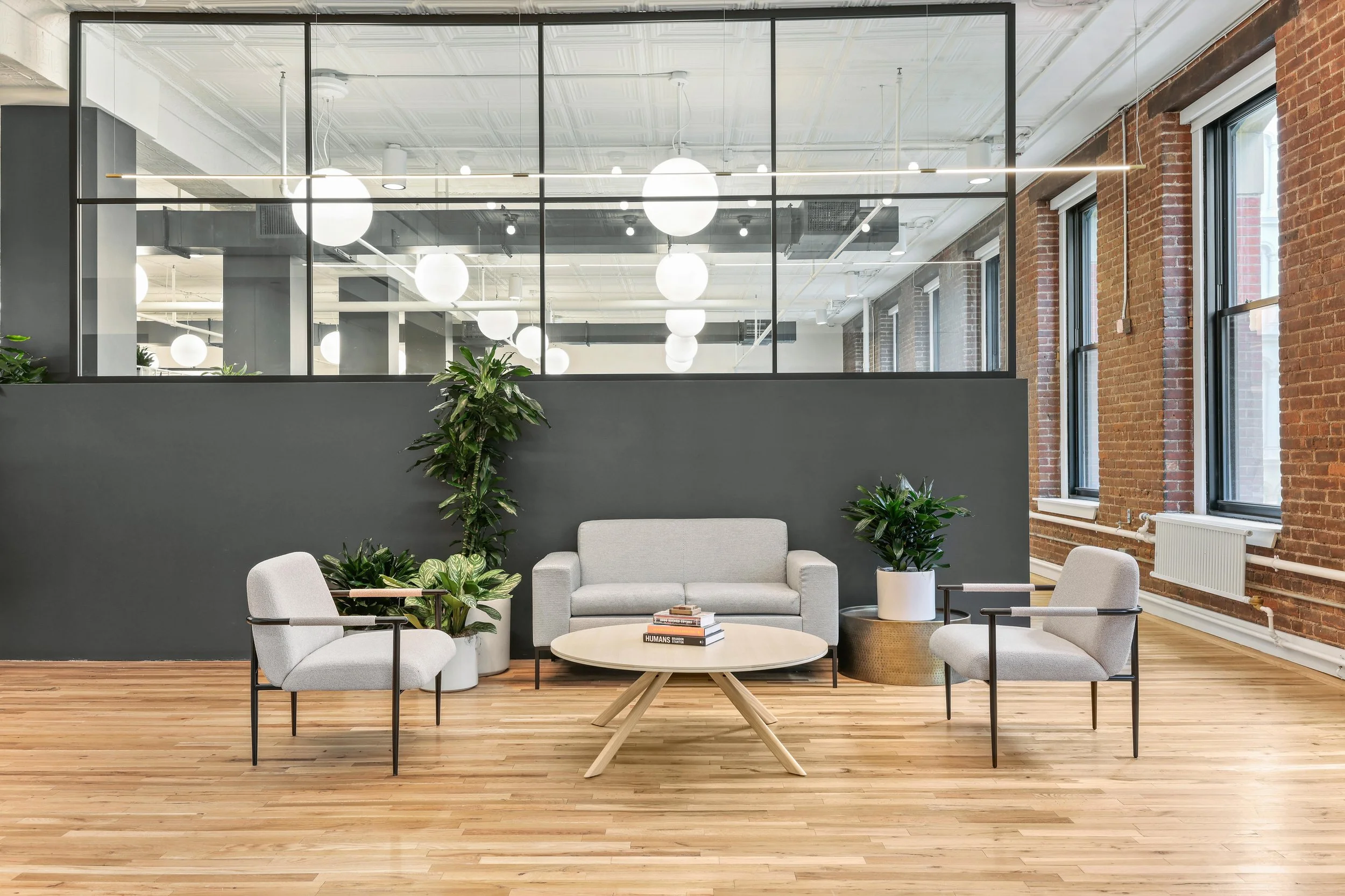 Modern office lounge area with a gray sofa, two matching armchairs, a round coffee table with books, and various potted plants on a wooden floor. Large windows with brick walls and glass partition above.