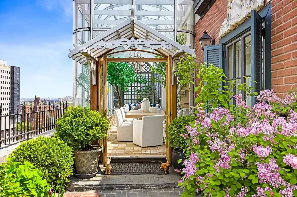 Rooftop garden patio with white seating area, lush greenery, pink flowers, brick and glass walls, and city view