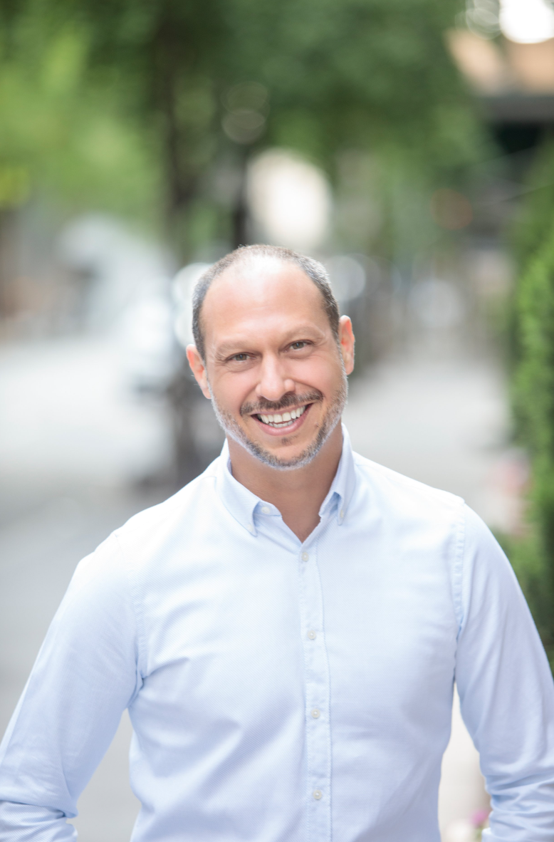 Smiling man with short hair and a beard wearing a white button-up shirt outdoors on a blurred street