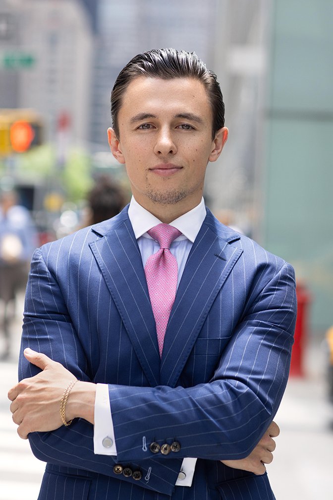 A young man dressed in a blue pinstripe suit with a pink tie, standing confidently outdoors in an urban setting with blurred cityscape background.