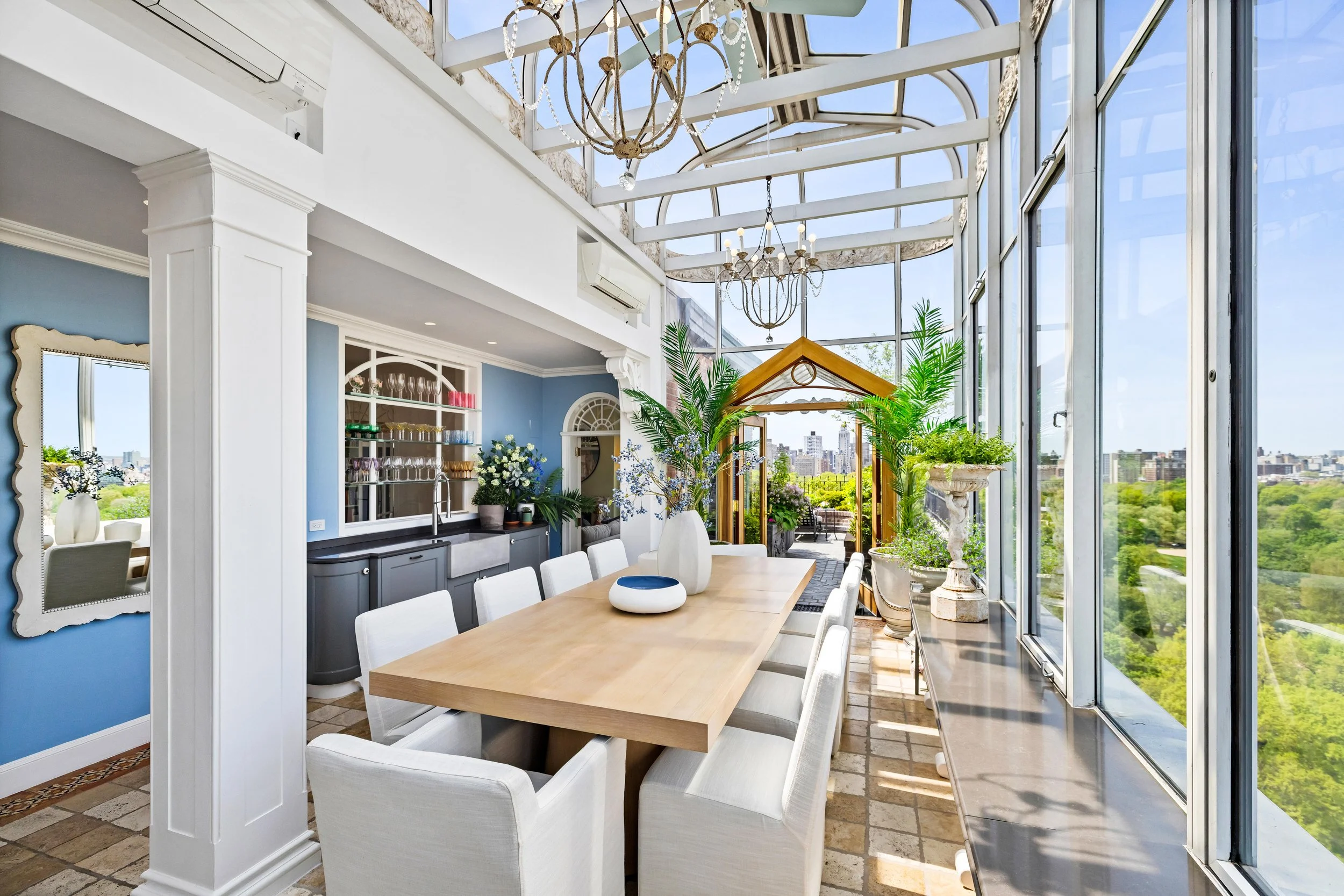 Sunlit dining area with large wooden table, white upholstered chairs, indoor plants, decorative vases, glassware, and a view of city skyline through glass walls and ceiling.