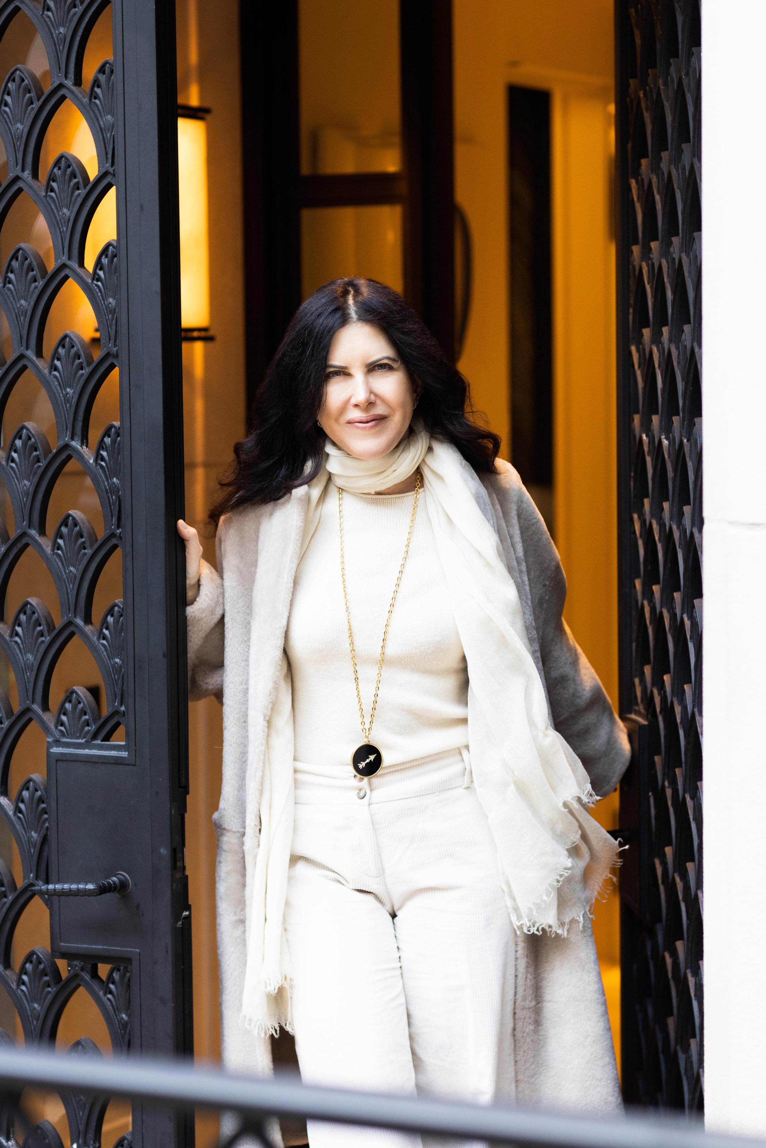 Woman with black hair smiling, wearing a beige outfit, standing in a doorway with black decorative gates, warm indoor lighting.
