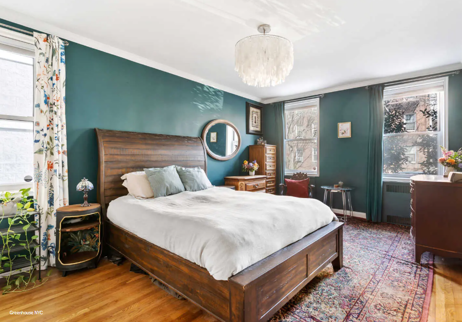 Bedroom with teal walls, large bed with white linens, wooden headboard, window with floral curtains, vintage furniture, and a chandelier.