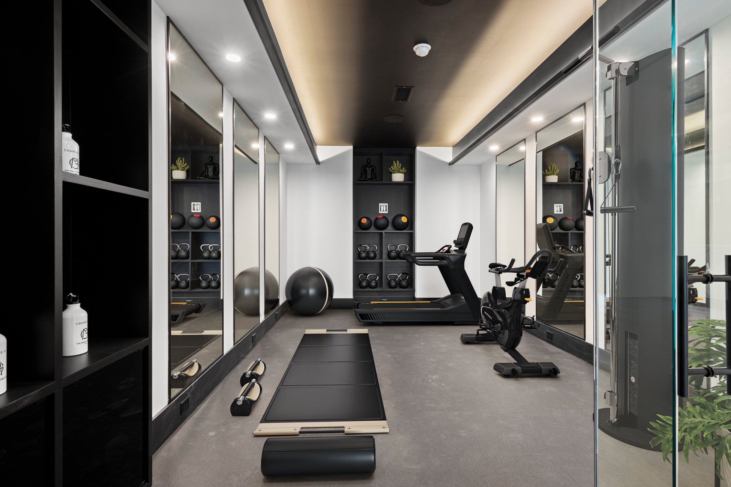 A modern, small home gym with treadmill, exercise bike, stability balls, and kettlebells, with mirrored walls and wall shelves holding decor and fitness equipment.