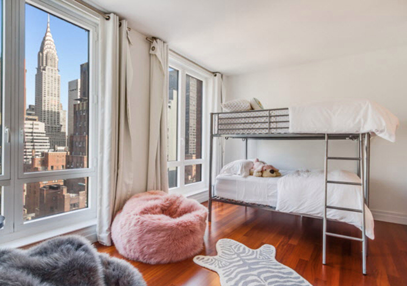 Bedroom with large windows showing cityscape view, bunk beds with white bedding, a pink furry chair, a gray fur rug, and a zebra print rug.