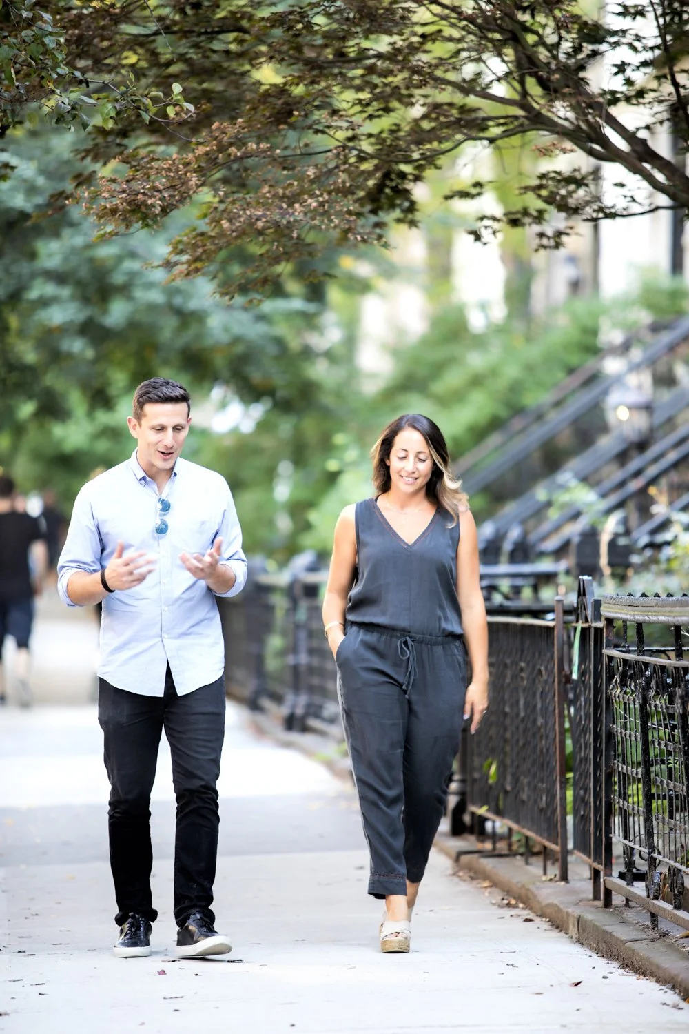 A man and woman walking together on a sidewalk, engaged in conversation. They are surrounded by greenery and trees, with a black metal fence on the side.