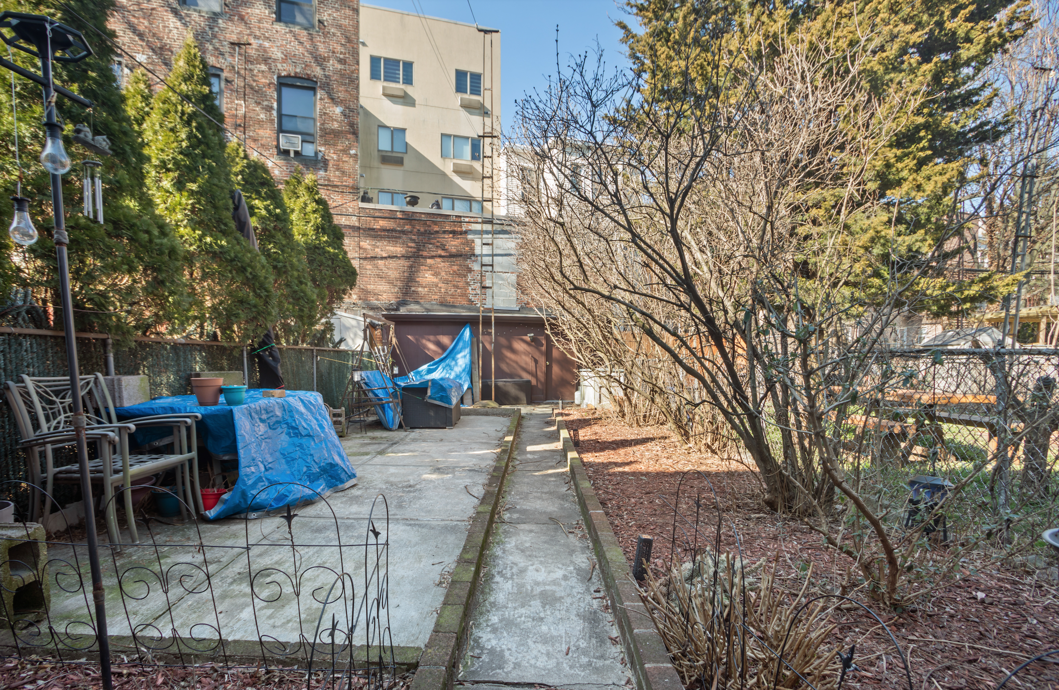 A backyard with a concrete patio, a blue tarp covering some objects, a small table with chairs, and a row of bare trees and bushes. Buildings are visible in the background.
