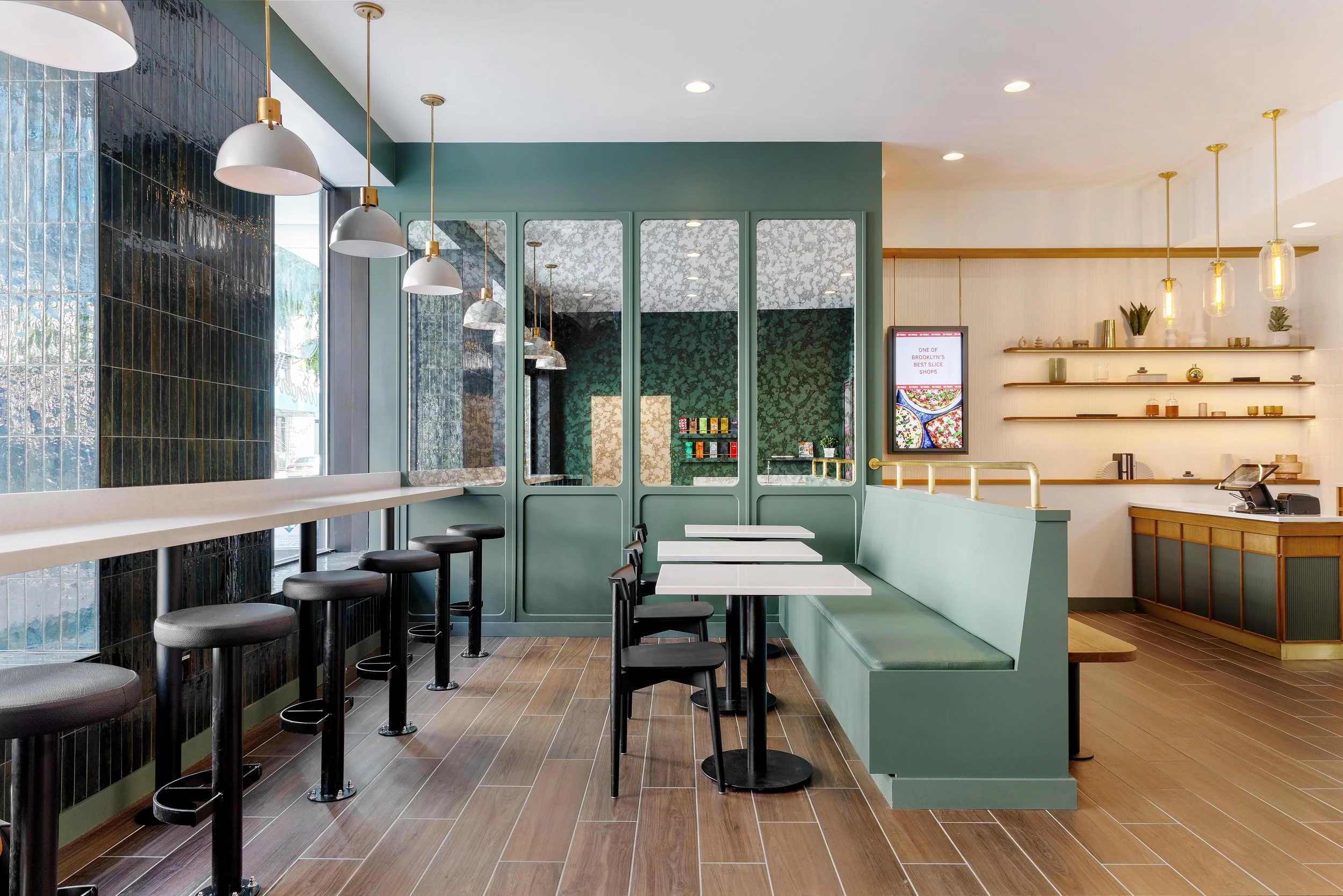 Interior of a modern cafe with a row of bar stools along a white counter on the left, a green seating area with white tables and black chairs in the center, and a white wall with floating shelves and a digital display on the right. There are hanging 