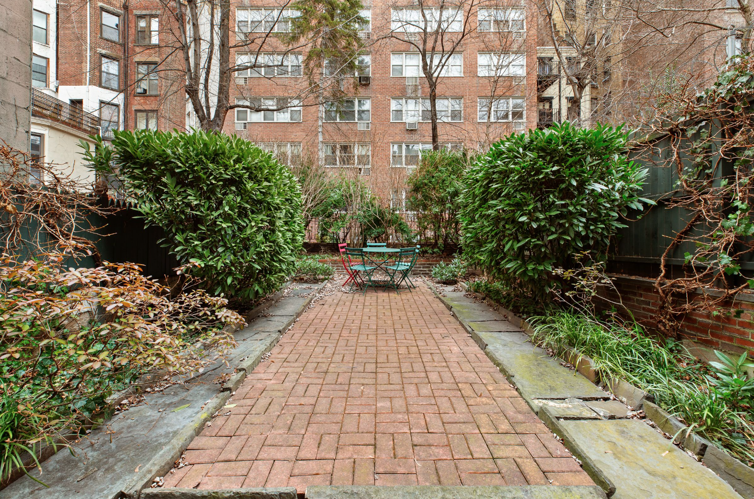 Small urban garden with brick pathway, two large bushes on each side, a round table with four colorful chairs in the center, surrounded by buildings and leafless trees.