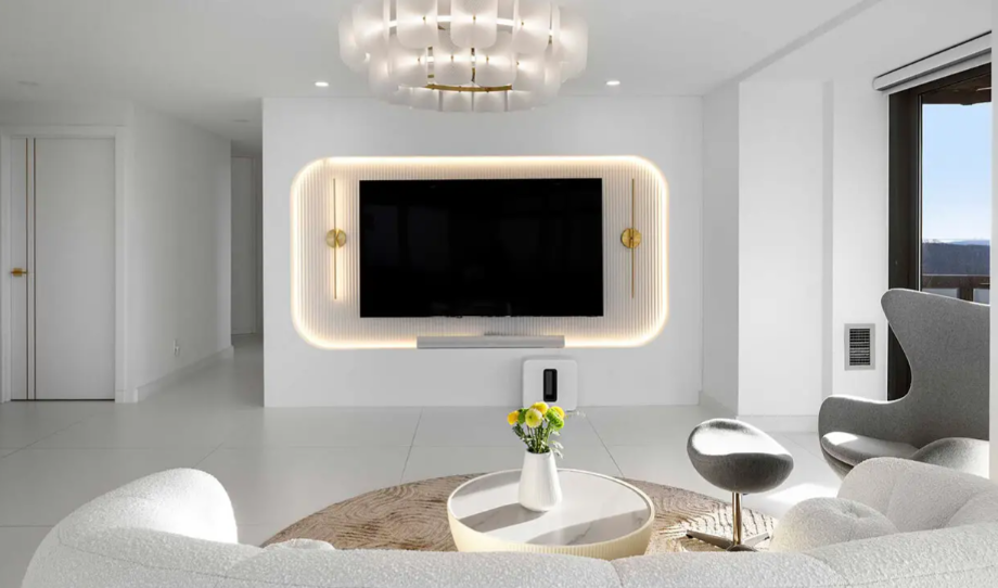 Modern living room with a wall-mounted flat-screen TV surrounded by a backlit frame, a round white coffee table with a vase of yellow flowers, and contemporary gray and white seating, with a large window showing an outdoor view.