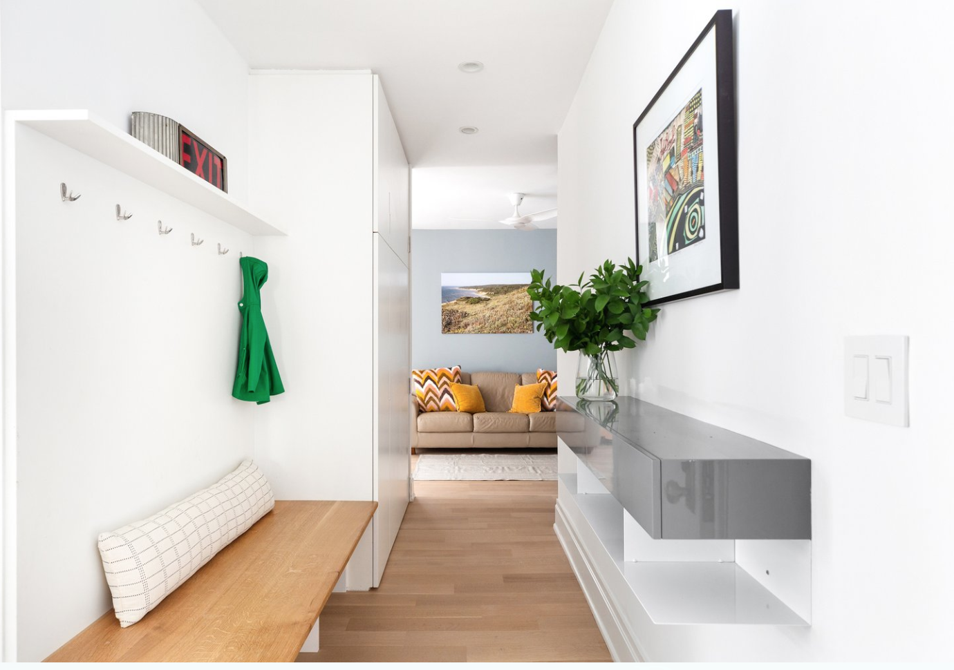 Modern entryway with white walls, a wooden bench, hooks with a green jacket hanging, a white shelf with an exit sign, a framed colorful artwork, a glass vase with greenery, and a view into a living room with a beige sofa and colorful pillows.