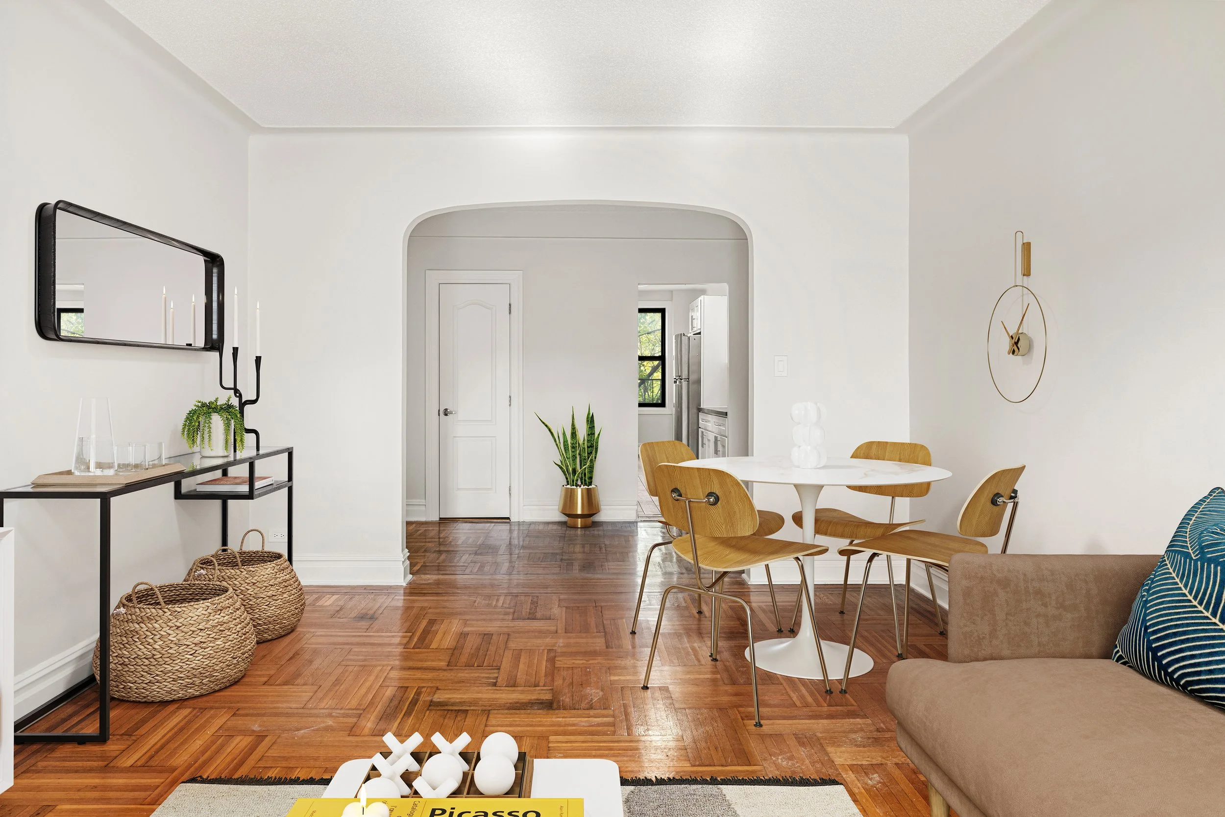 Living and dining room with white walls, hardwood flooring, a beige sofa with a blue patterned pillow, a white round dining table with four wooden chairs, a wall clock, a black console table with decorative items, a wall mirror, a potted plant, and a