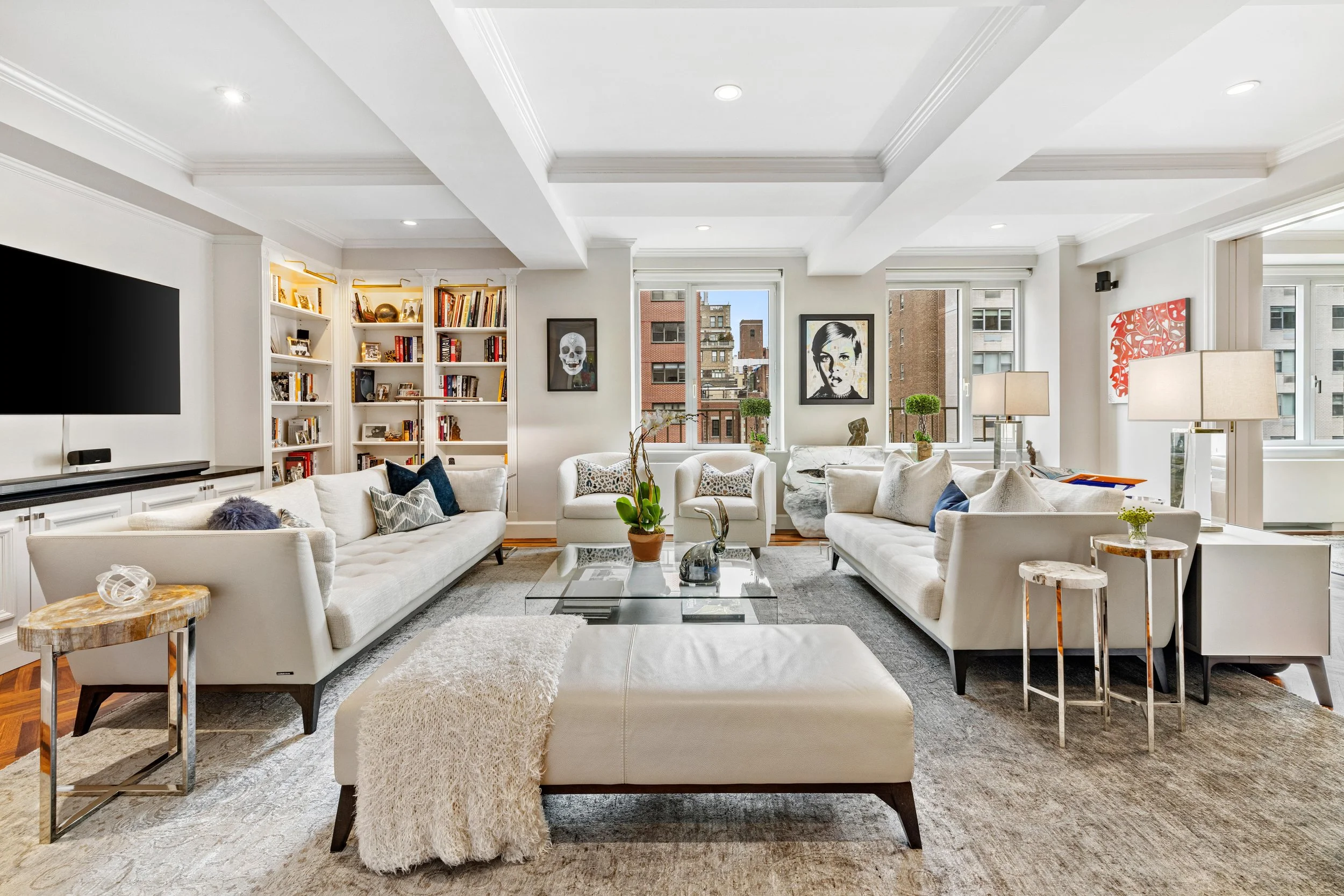 Bright and modern living room with white sofas, a glass coffee table, artwork on the walls, and large windows showing city buildings.