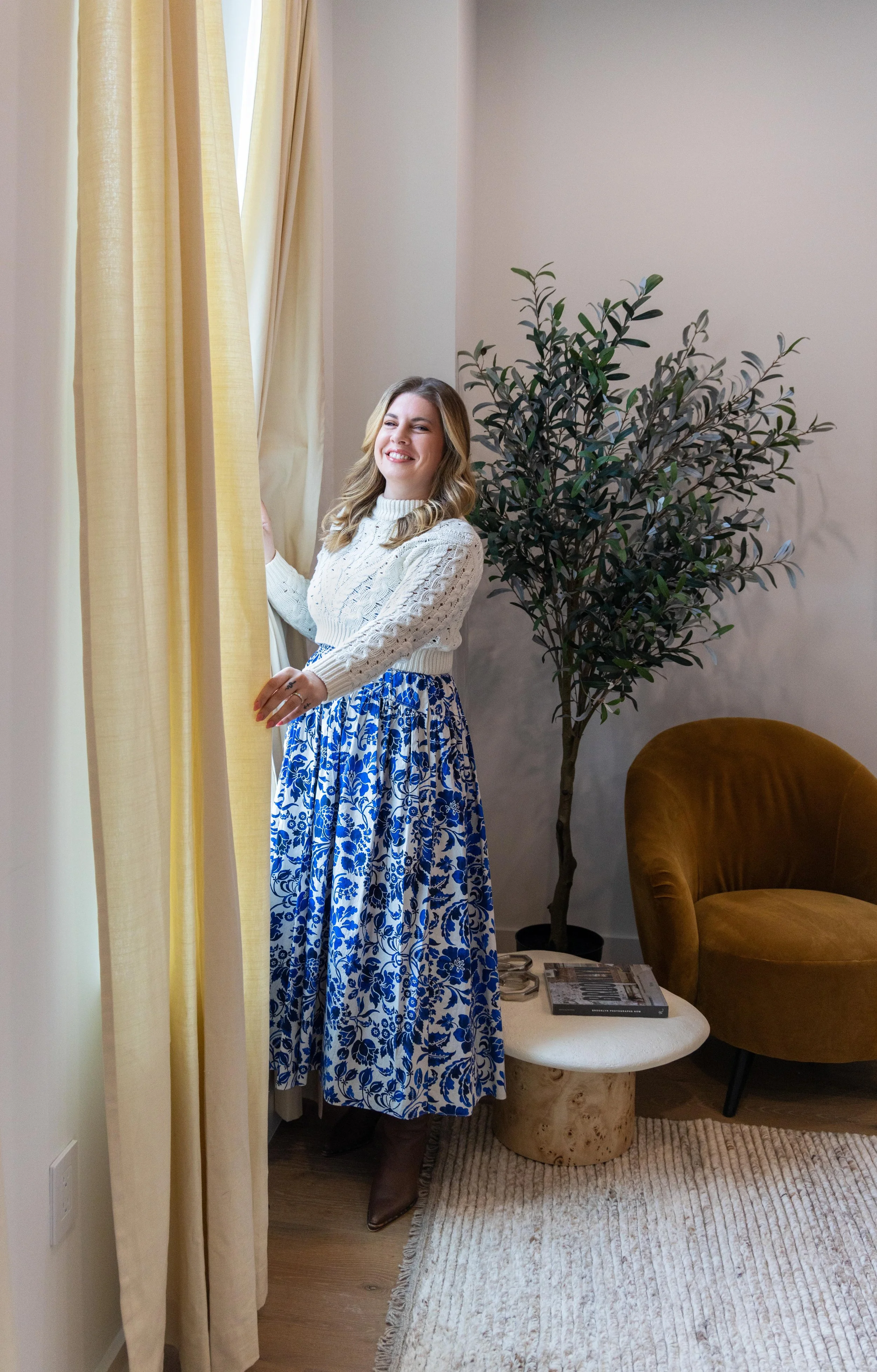 A woman with blonde hair, wearing a white cable-knit sweater and a blue and white floral long skirt, standing near yellow curtains in a cozy living room.