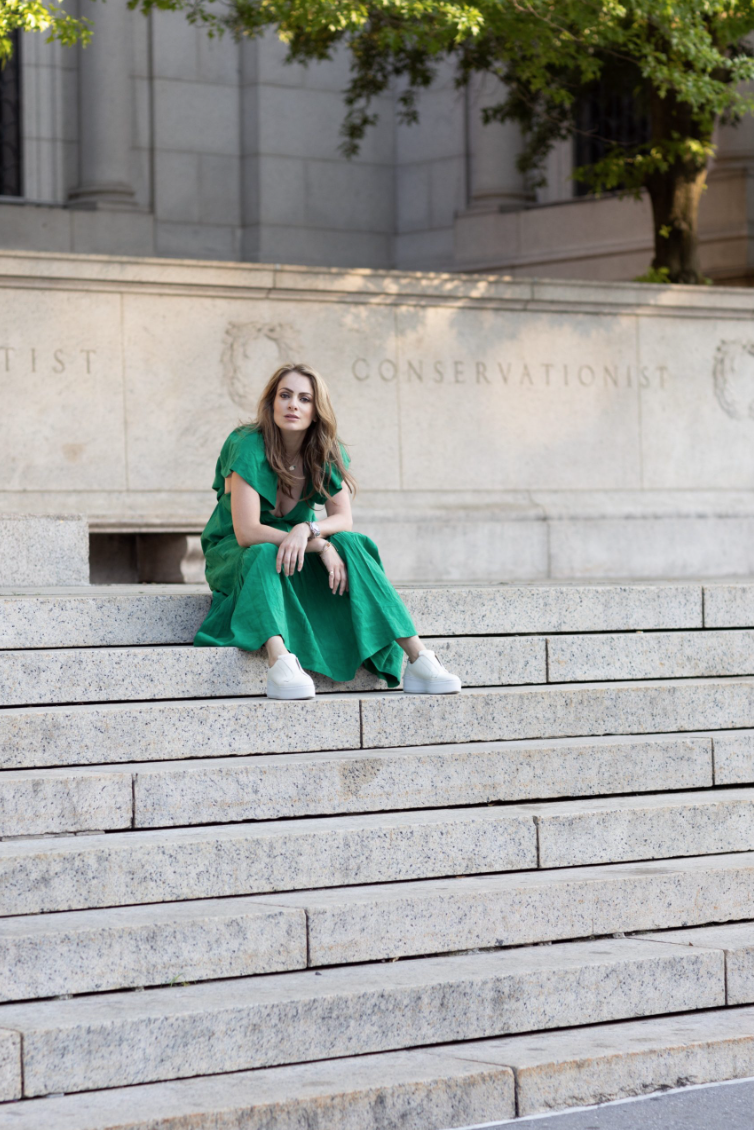 A woman in a green dress and white sneakers sitting on stone steps outside a building with trees and a conservationist inscription.