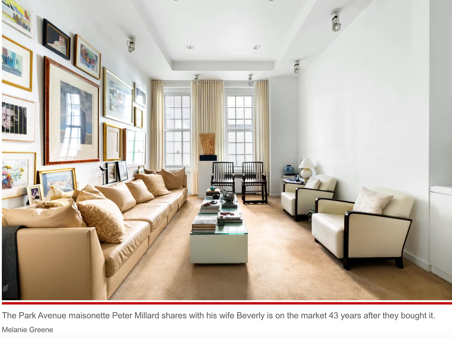 A bright living room with a long beige sofa and cushions, two armchairs, a coffee table with books and decorative items, large windows with white curtains, and artwork on the wall.