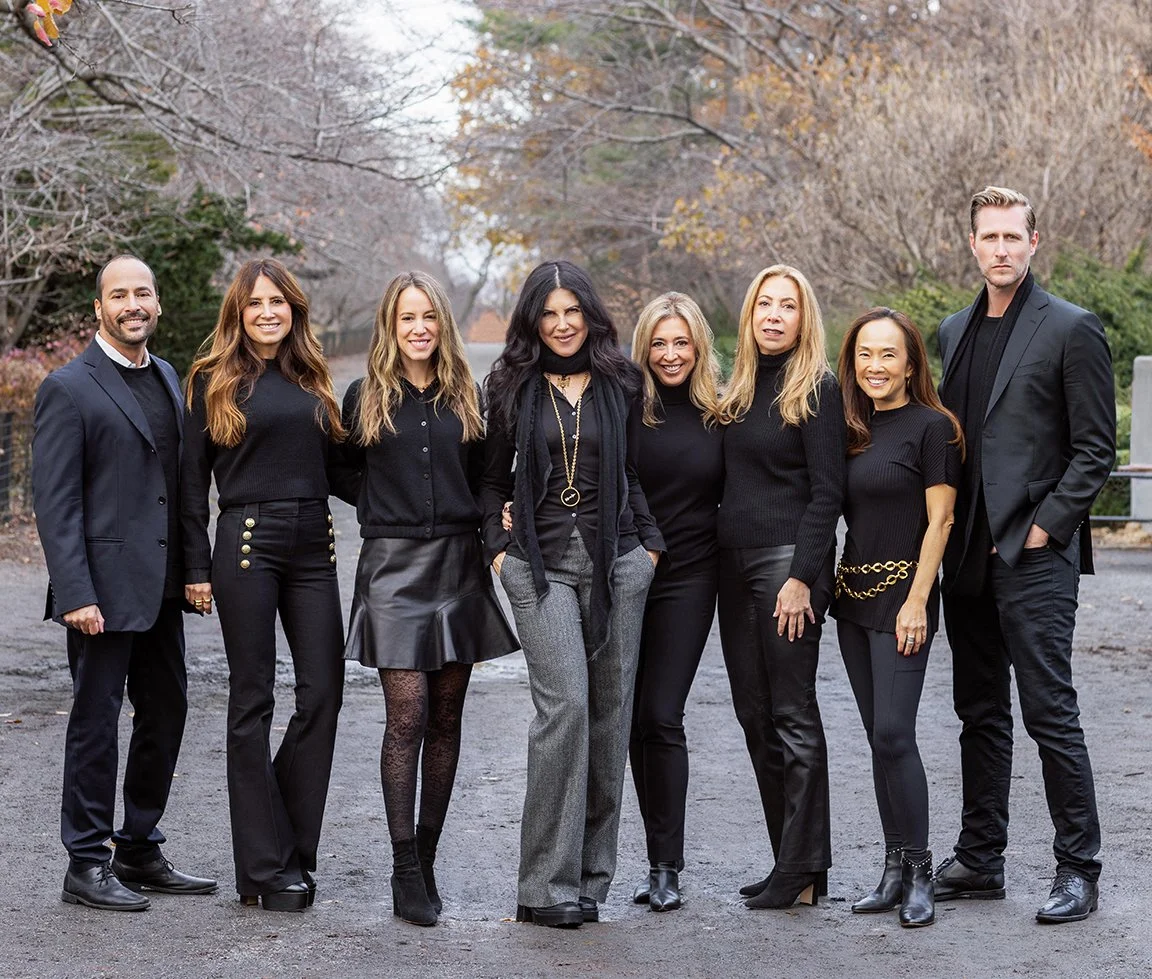 Group of nine diverse people standing outdoors in fall, dressed in black and gray attire, smiling for a photo.