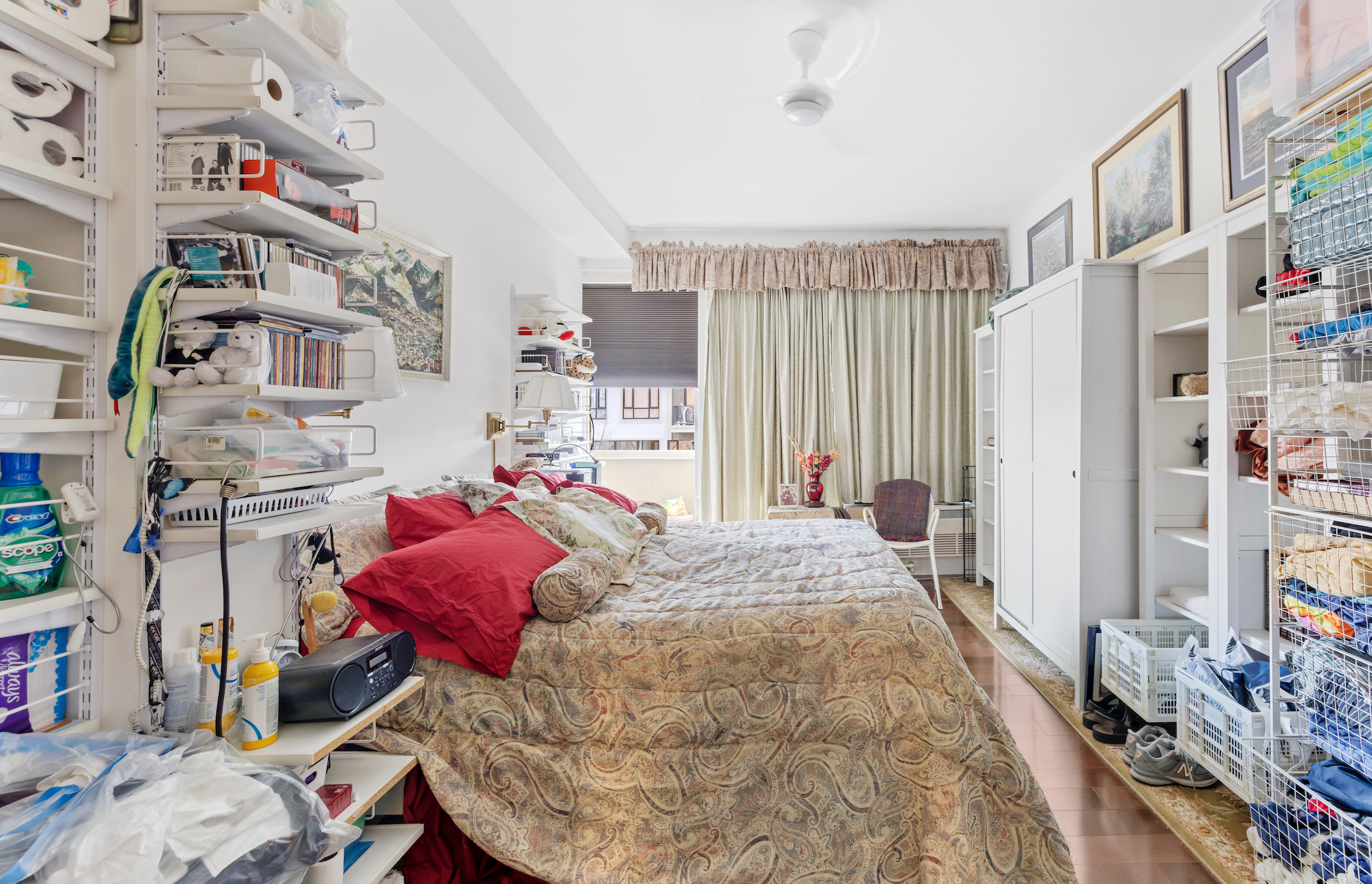 A cluttered bedroom with a bed featuring a paisley bedspread and multiple pillows, including red and patterned ones. The bed is against a white wall with shelves stocked with various items like books, toiletries, and miscellaneous objects. To the right, there are white shelving units and a wire storage rack filled with clothes and fabric. Near the window, there's a small table with a red vase and flowers, a chair, and an overall cozy but untidy atmosphere with curtains and a ceiling fan.