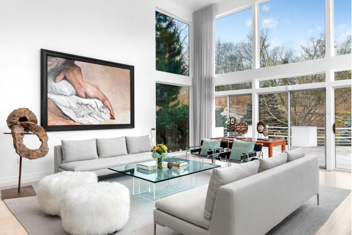 Bright, modern living room with floor-to-ceiling windows, white walls, light-colored furniture, a glass coffee table, artwork, and decorative sculptures.