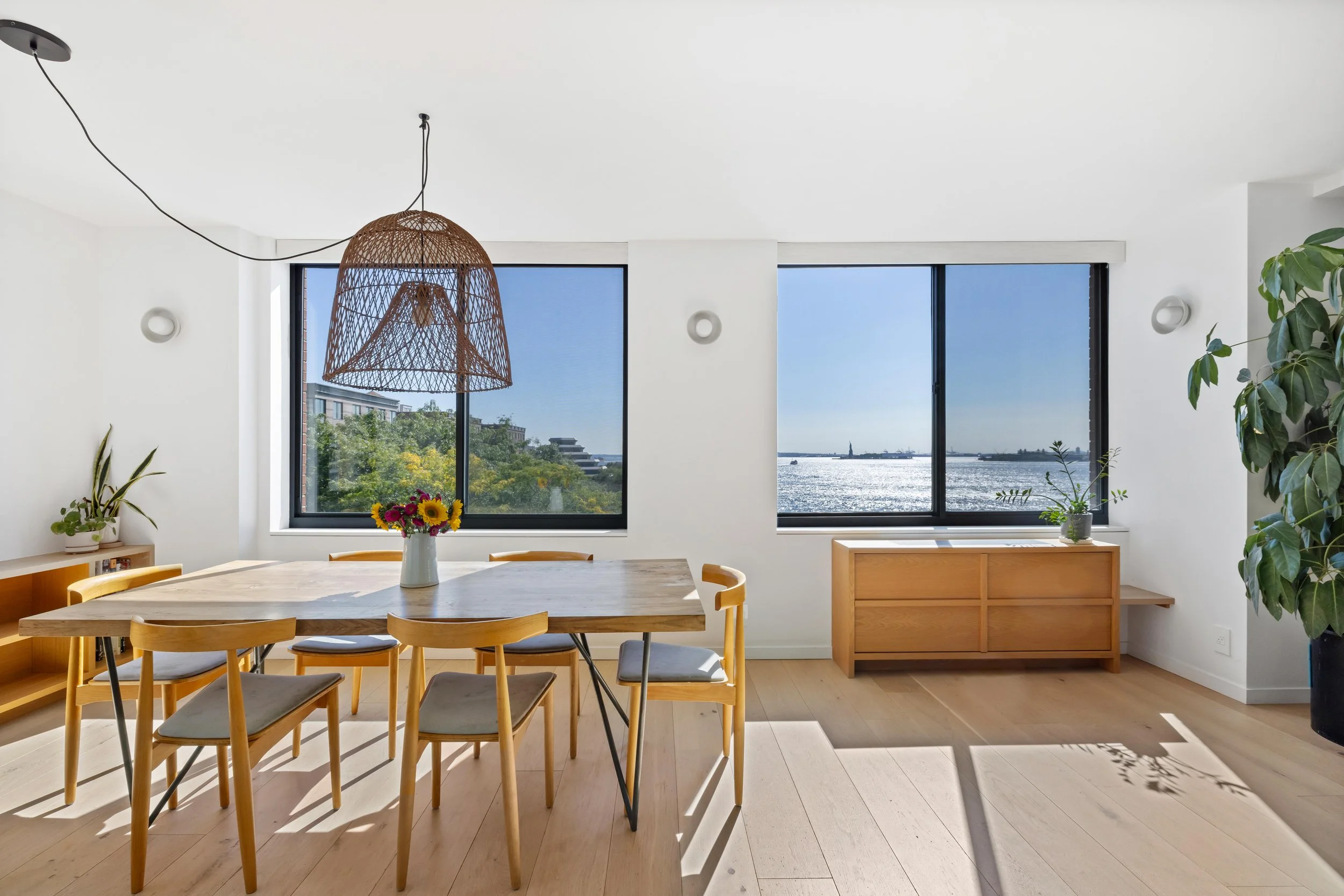 Bright dining room with large windows showing water view, wooden dining table with six chairs, hanging wicker light fixture, and potted plants.