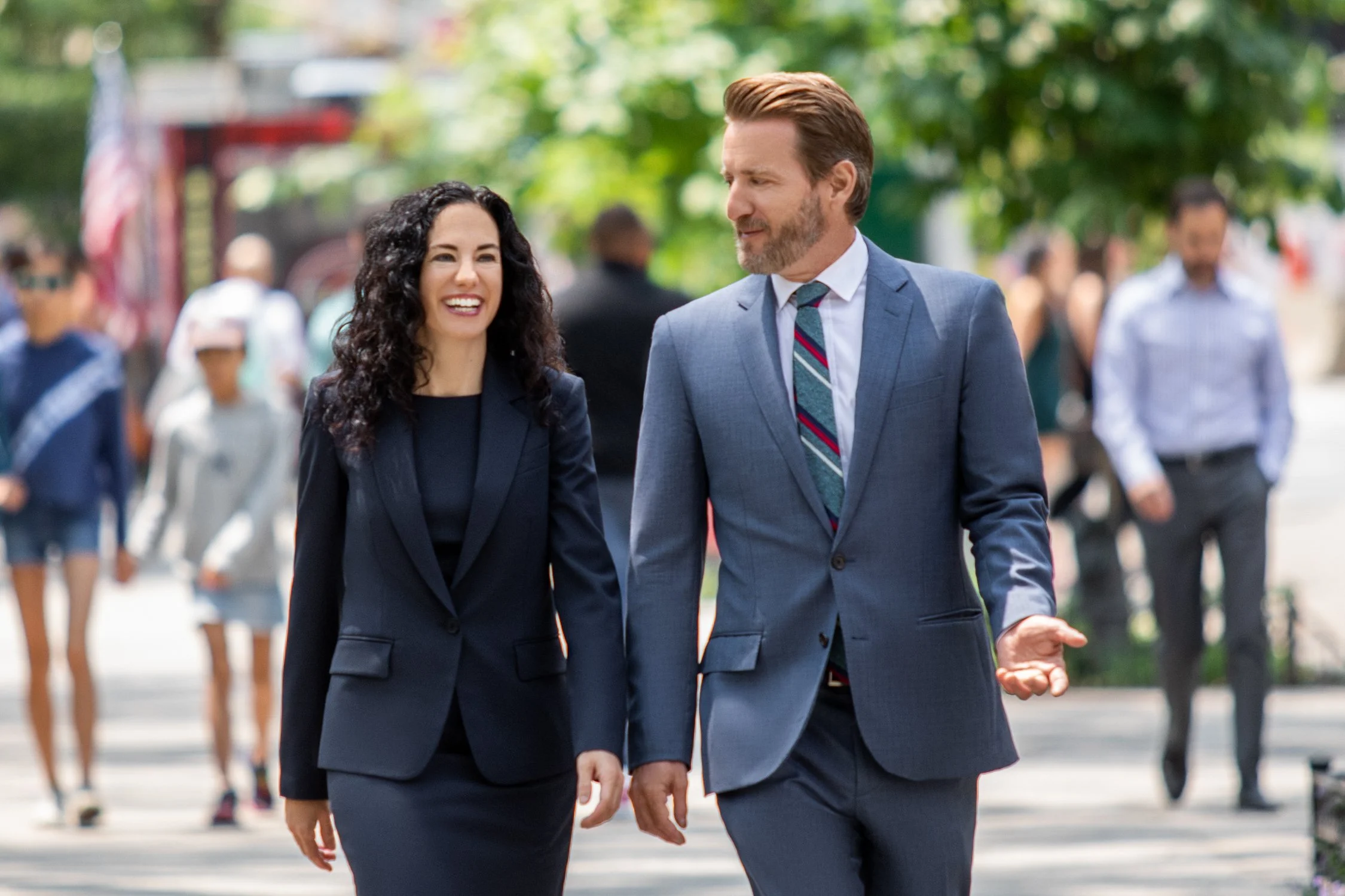 A man and woman walking together outdoors, dressed in business attire, smiling and talking, with blurred pedestrians and trees in the background.