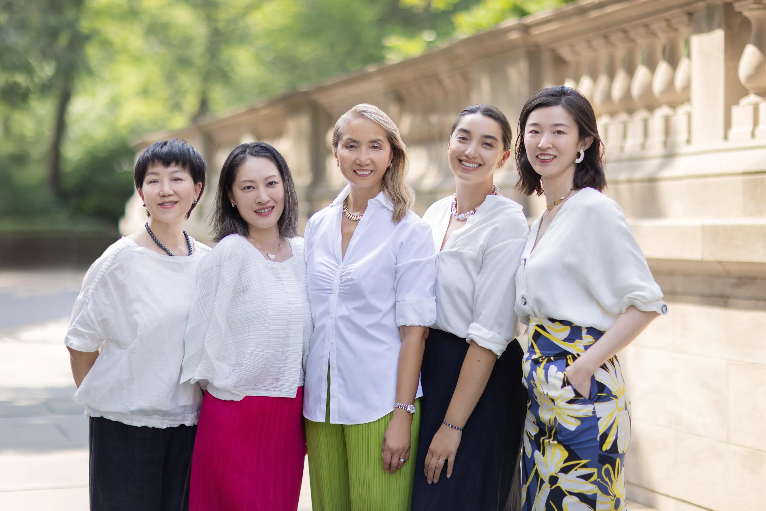 Group of five women standing outdoors on a sunny day, dressed in casual and semi-formal white tops, smiling, with a stone balustrade and green trees in the background.