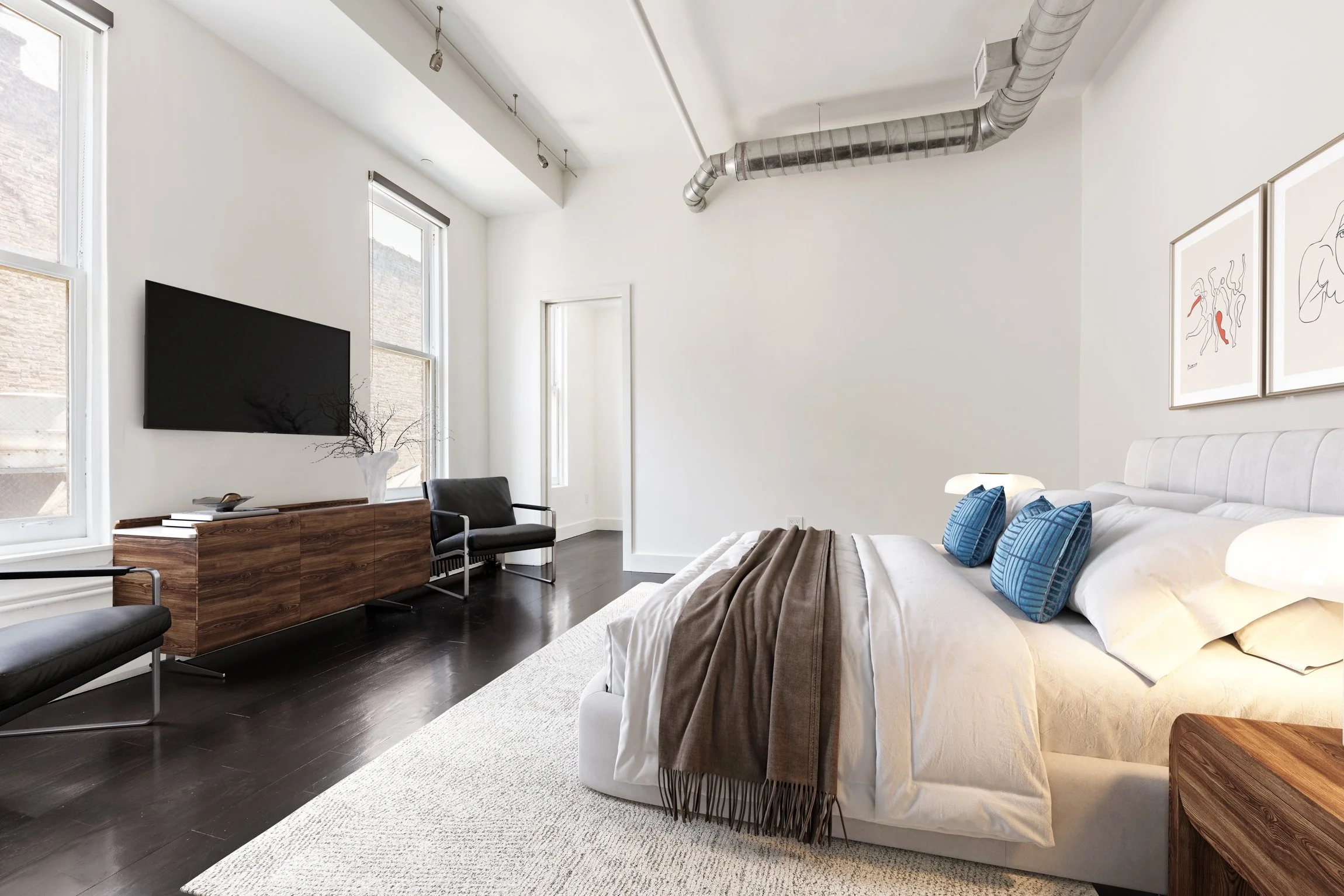 Modern bedroom with a white bed, blue pillows, brown throw blanket, dark wood nightstand, black chairs, wooden TV stand, mounted flat-screen TV, large windows, and minimalist artwork on the wall.
