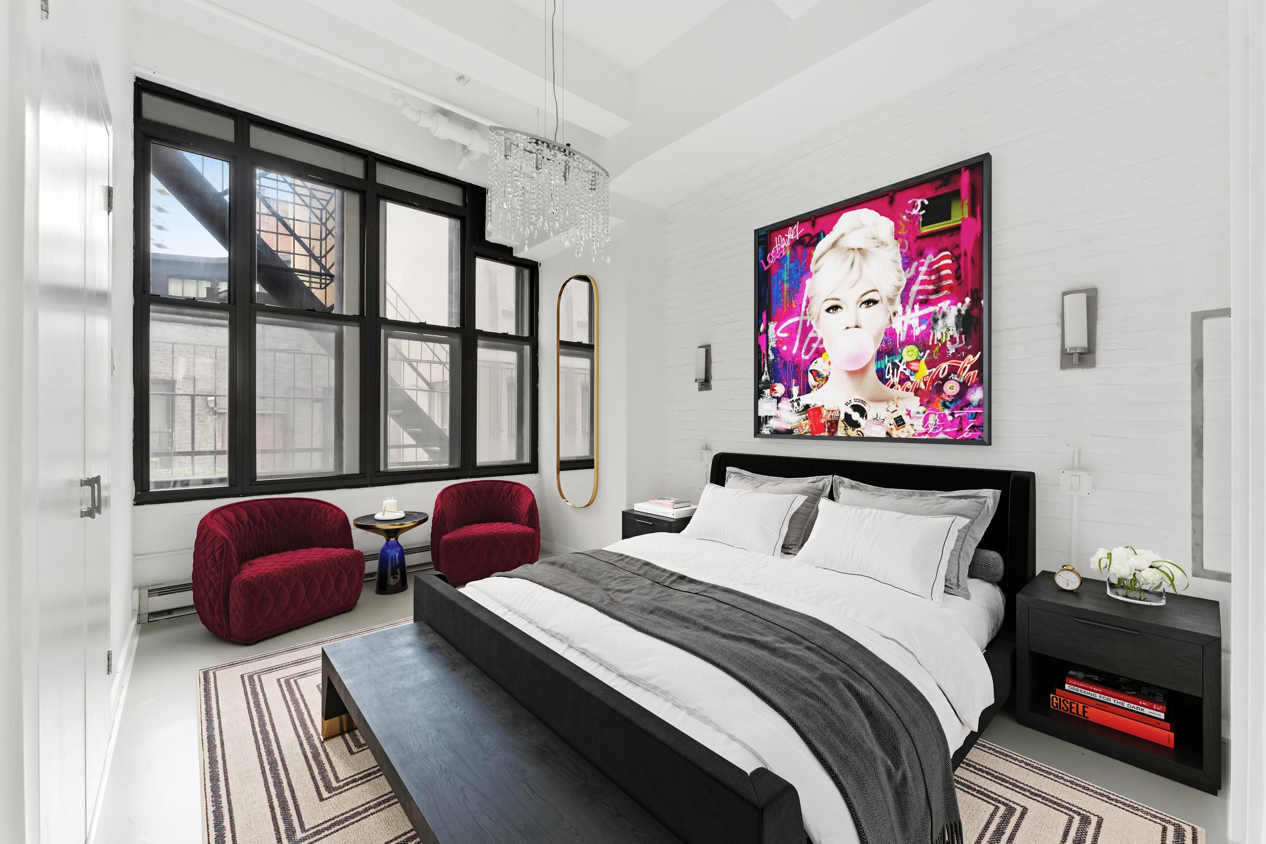 Modern bedroom with a black bed frame, white bedding with gray accents, black nightstand with books, white walls, large window with black frames, red velvet chairs, round side table, chandelier, abstract art on wall, mirror, clock, and decorative pla
