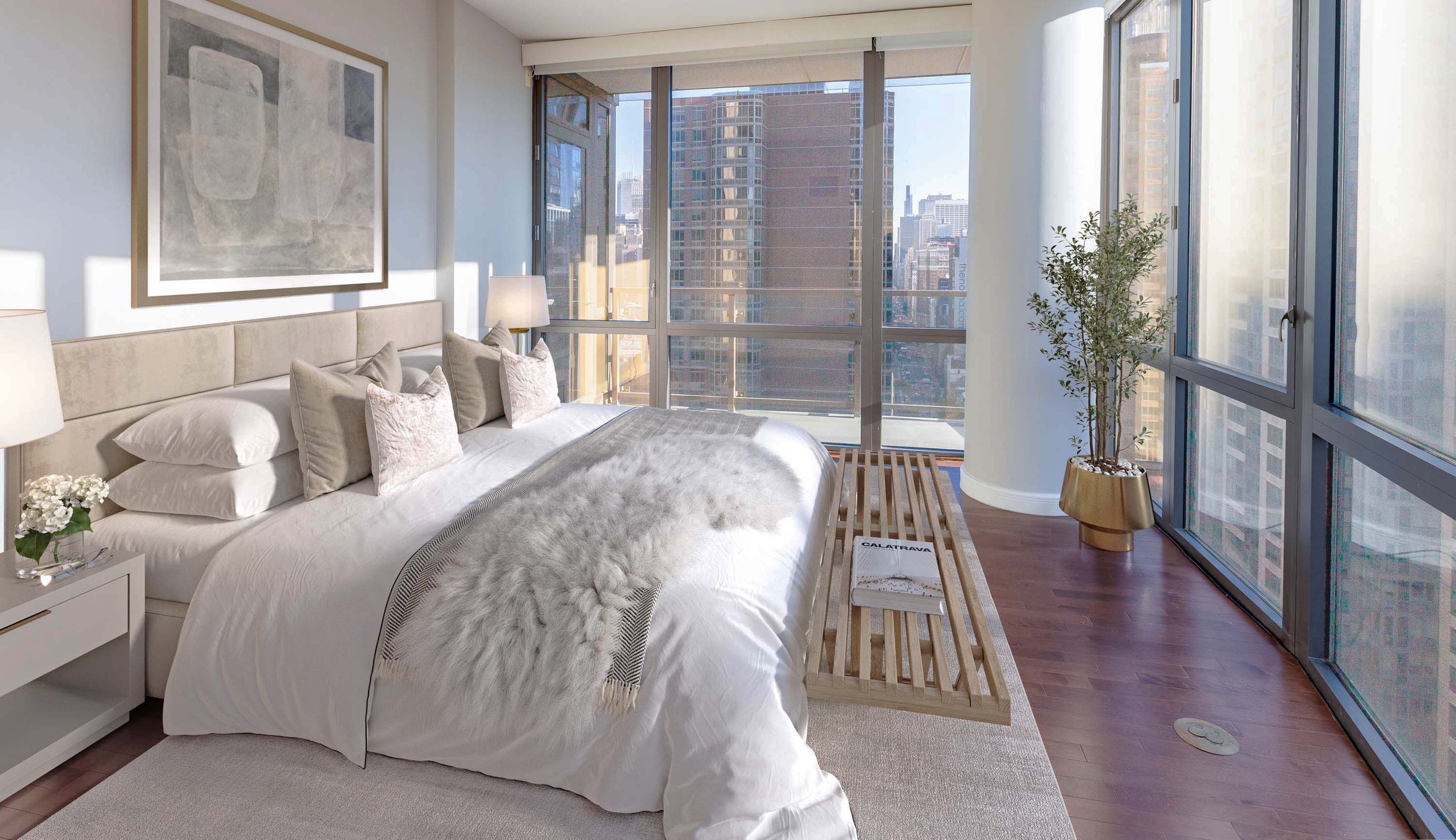 Modern bedroom with large windows, a white bed with multiple pillows, a nightstand with flowers and lamps, a wooden bench, a potted plant, and cityscape view.