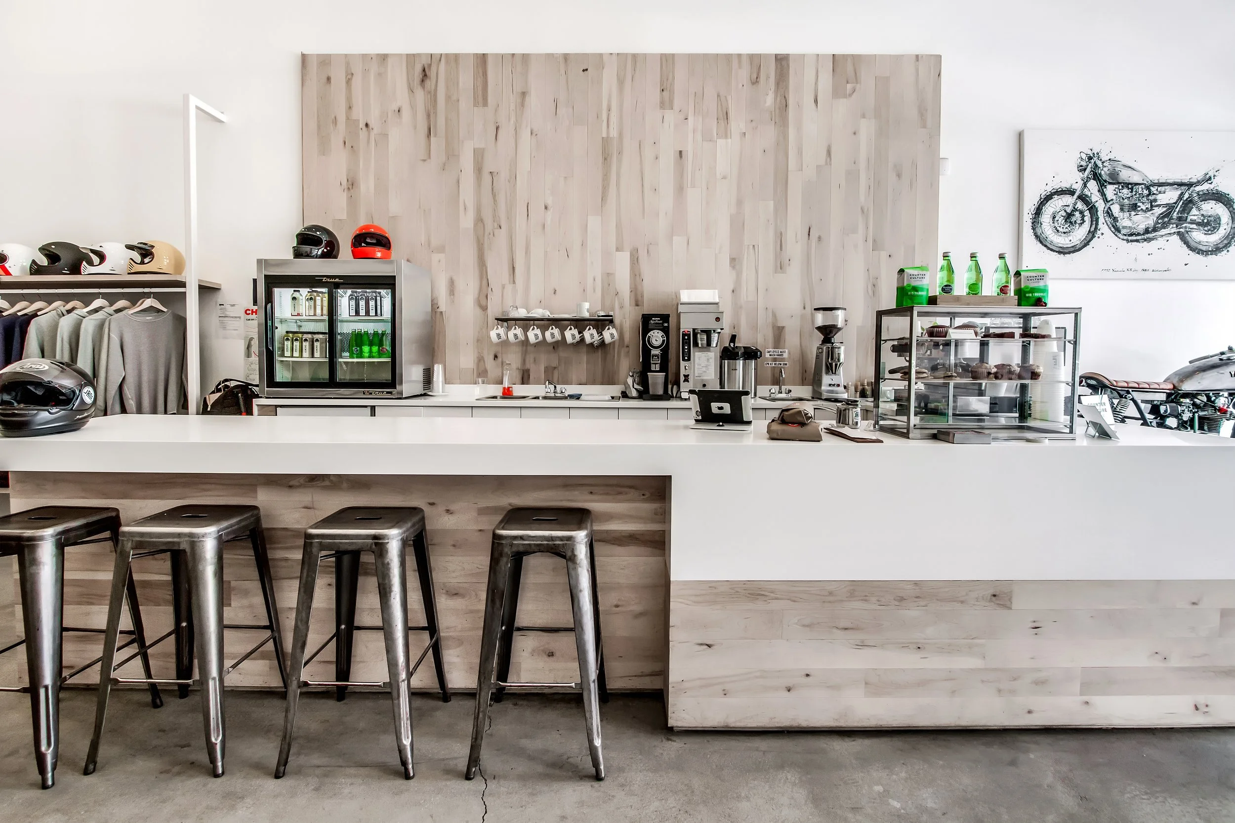 A modern coffee shop counter with four metal bar stools, a wooden back wall, a display case with baked goods, and various coffee-making equipment and bottles.