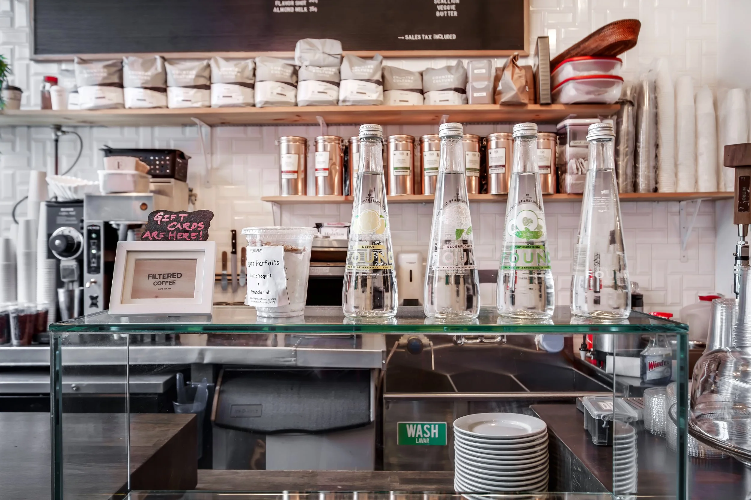 A coffee shop counter with four bottles of flavored sparkling water, a small sign indicating gift cards are available, a container of yogurt parfaits, a glass display case, and shelves with coffee beans and supplies in the background.