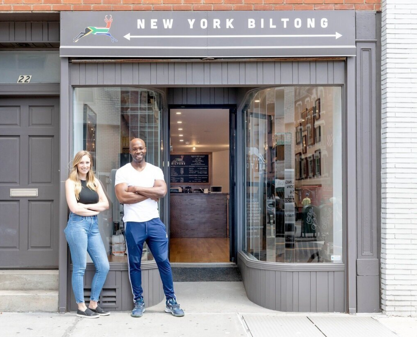 Two smiling people standing outside a restaurant with a sign that reads "New York Biltong."