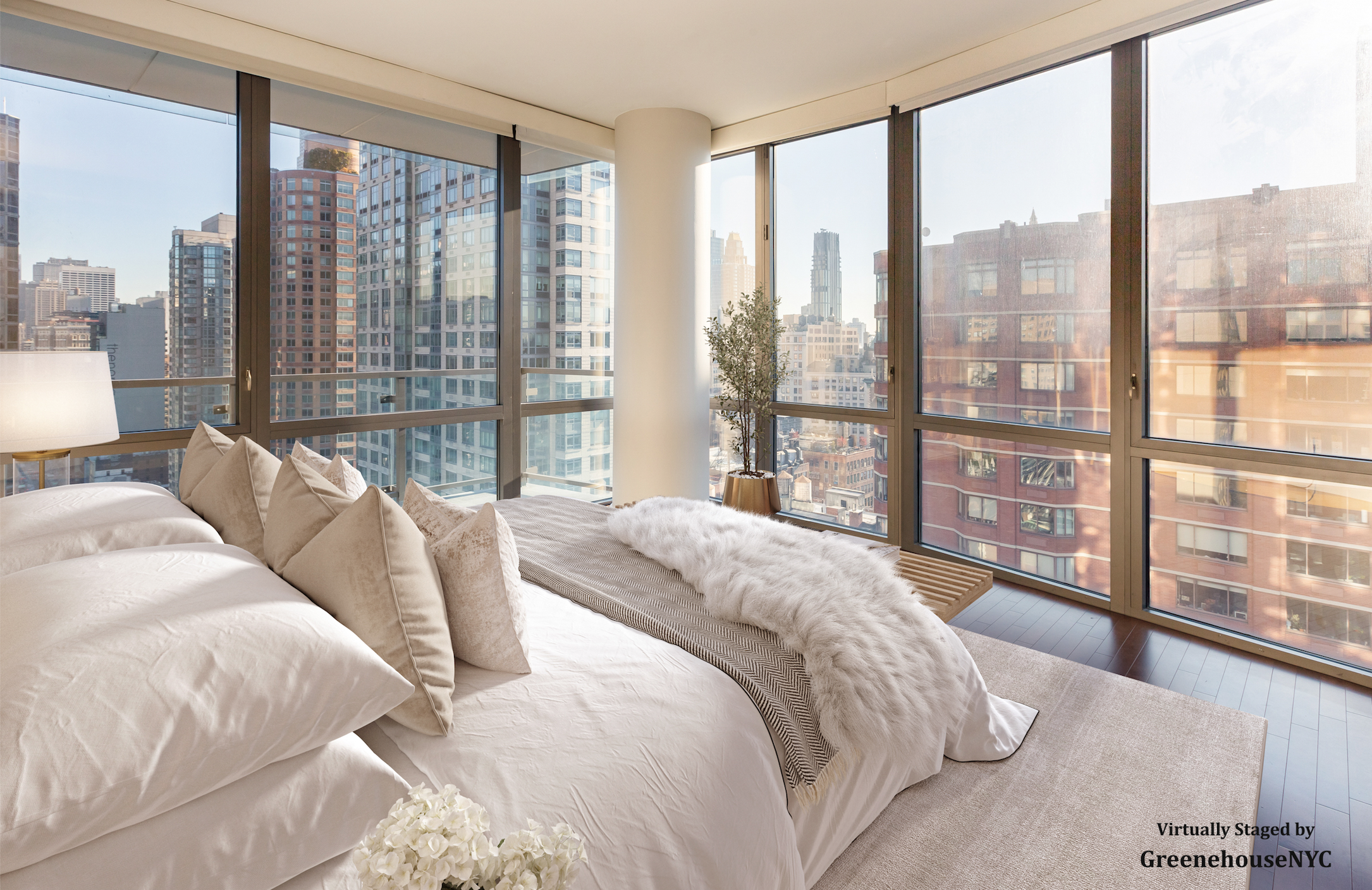 Luxurious bedroom with large windows offering city views, a bed with multiple beige pillows, a white duvet, a fuzzy throw, and a small flower arrangement near the bed.