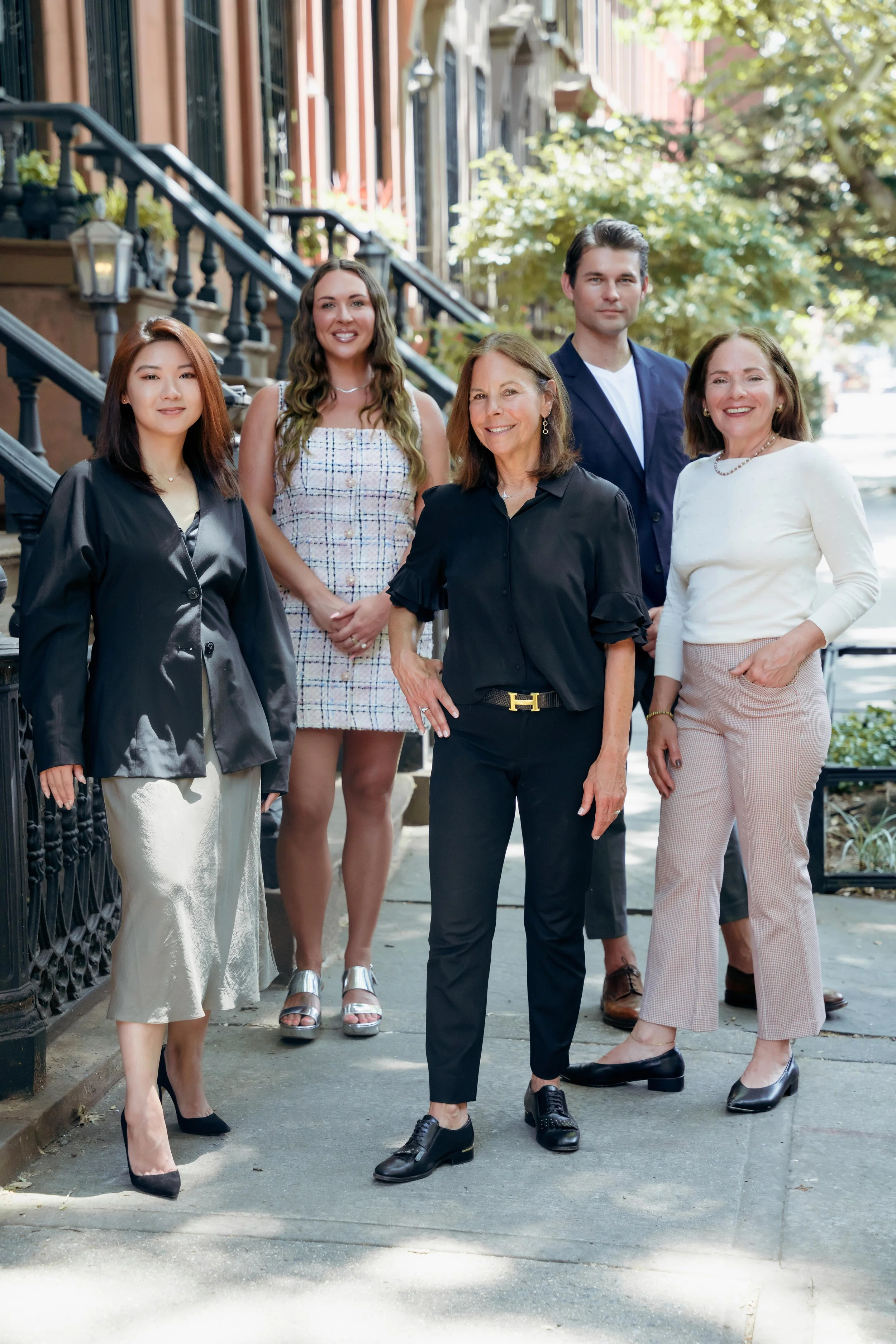 Group of five professionally dressed women and one man standing on a city sidewalk in front of brownstone buildings and trees.