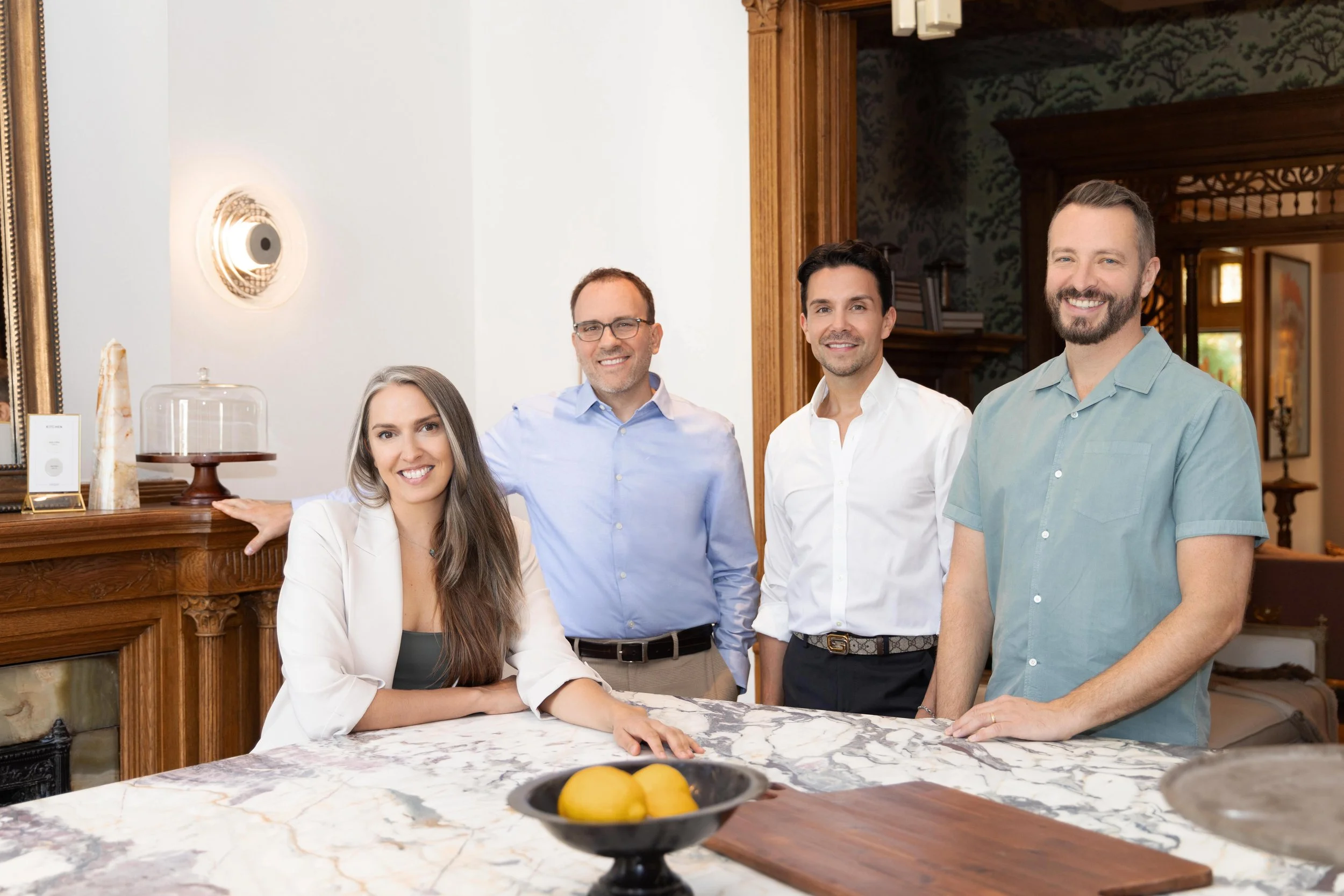 Four people, two men and two women, smiling and posing in a kitchen with a marble countertop, wooden cabinetry, and decorative wall art.