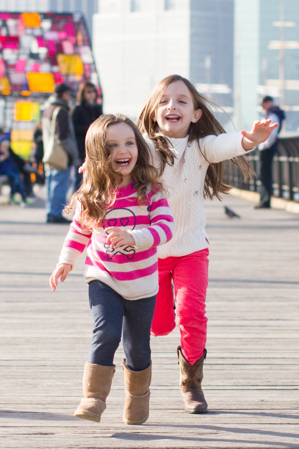 Two young girls running and smiling on a wooden pier, with colorful blurred objects and people in the background.