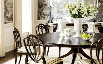 A dining room with a round wooden table, six chairs with patterned cushions, a vase of white flowers, and a window with curtains in the background.