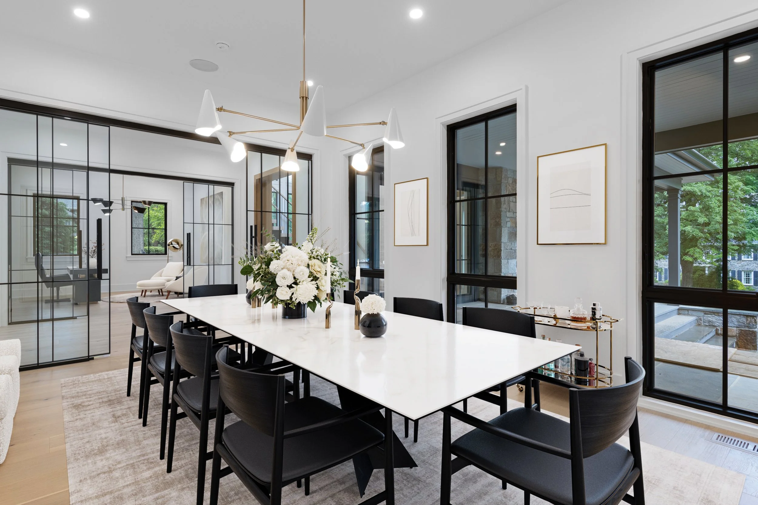 Modern dining room with a white rectangular table, black chairs, floral centerpiece, large windows, and minimal art on white walls.