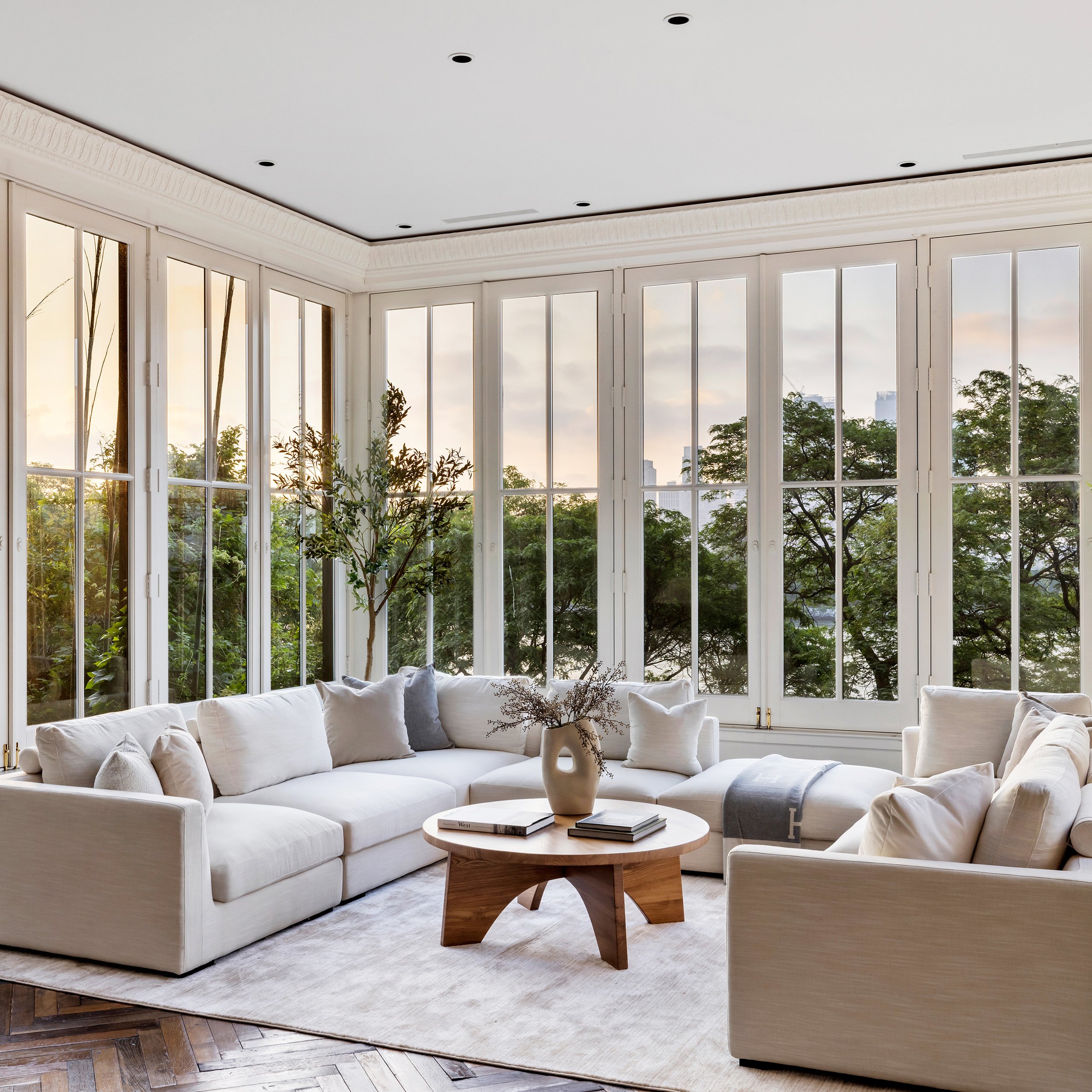 Bright living room with large windows, white sectional sofa with pillows, a wooden coffee table with books and a vase, and greenery outside