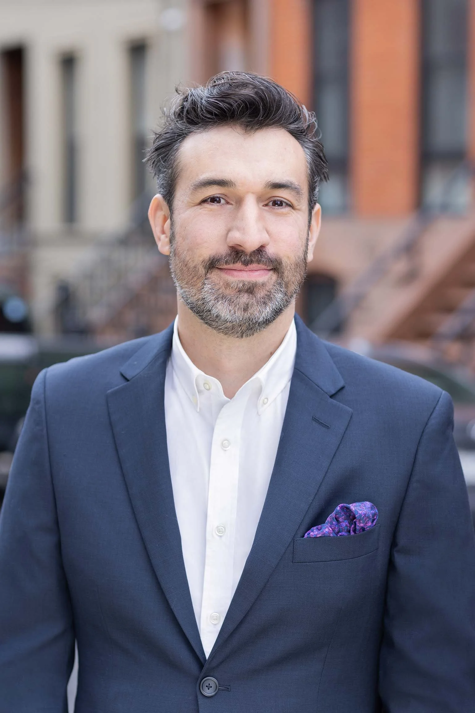 A middle-aged man with dark hair and a beard, wearing a navy suit jacket and white dress shirt, standing outdoors in an urban setting with brick buildings in the background.