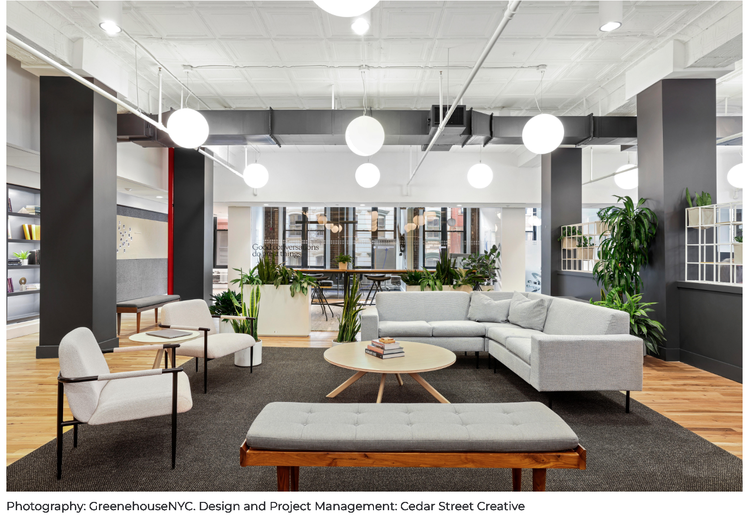 Modern coworking space with lounge seating, potted plants, books, and large windows allowing natural light.