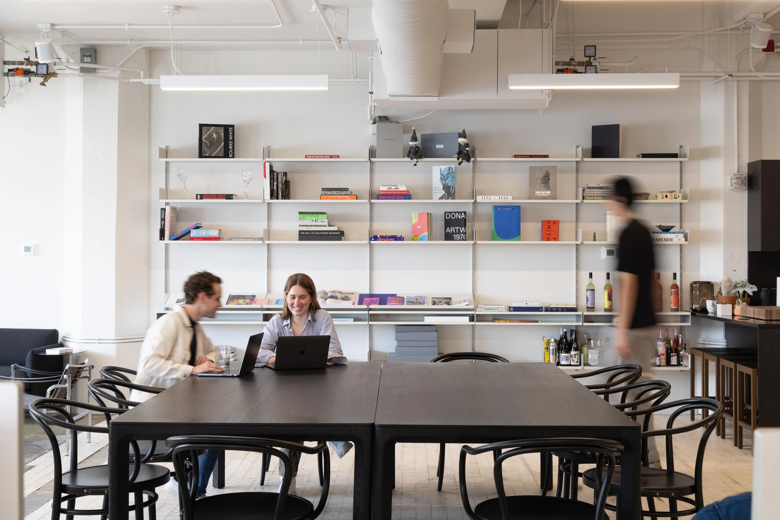 Two women working on laptops at a large black table in a bright, modern room with white walls and a bookshelf behind them. One man is walking past, blurred in motion.