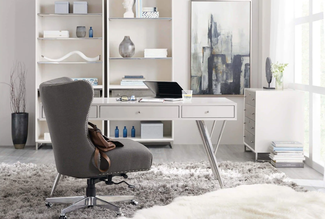 Modern home office with white desk, gray office chair, white bookshelves, large abstract painting, and natural light from windows.