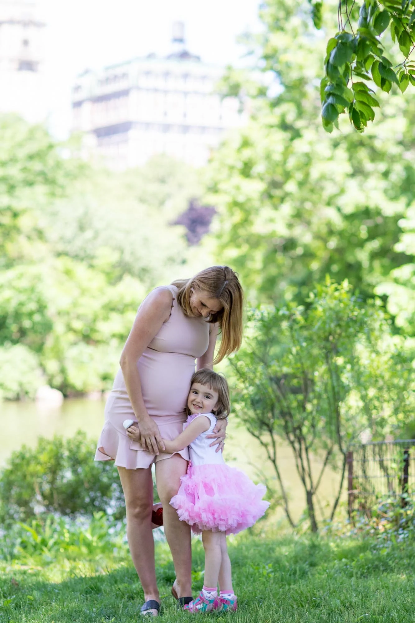 A woman in a light pink dress is hugging a young girl in a pink tutu dress outdoors in a lush green park with trees and water in the background.